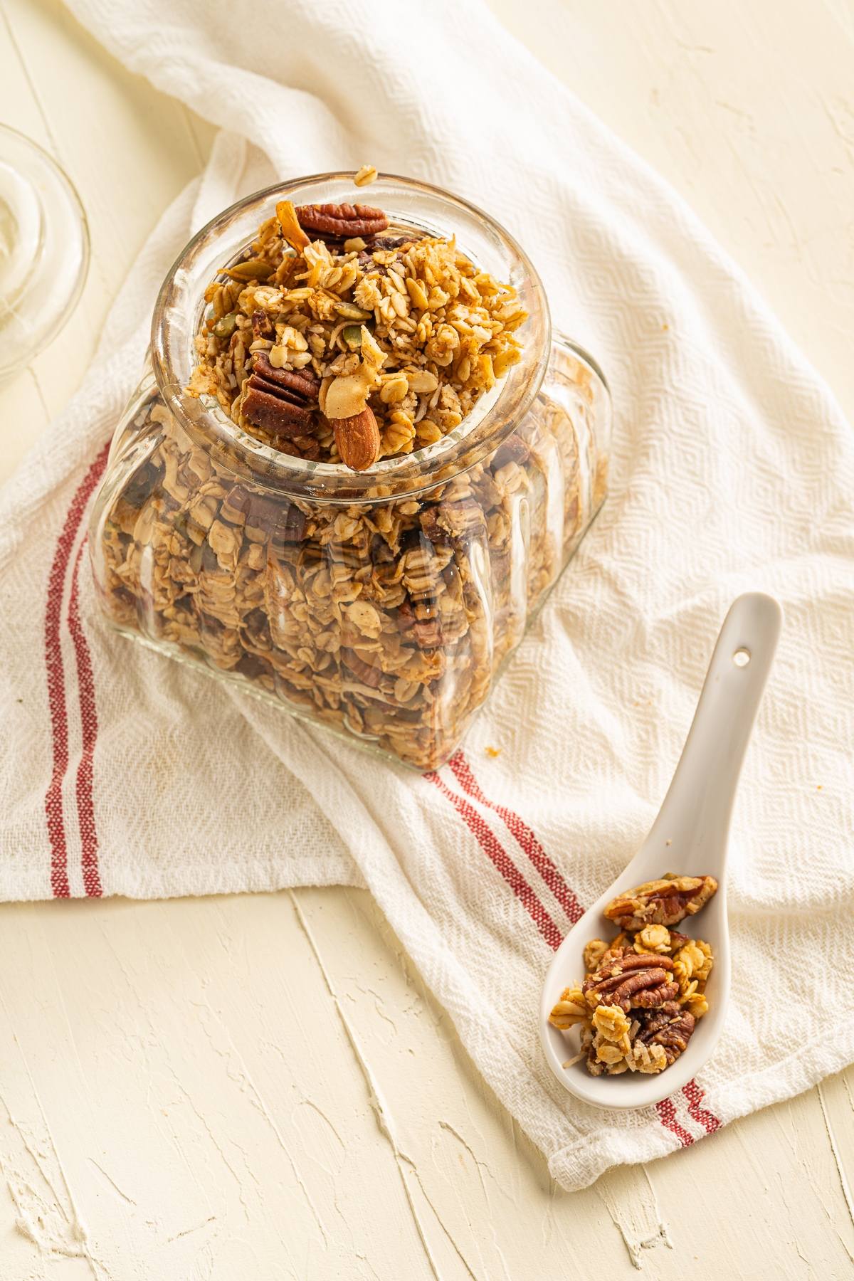 A glass jar of easy homemade granola with nuts sits on a towel, with a spoonful of granola beside it.