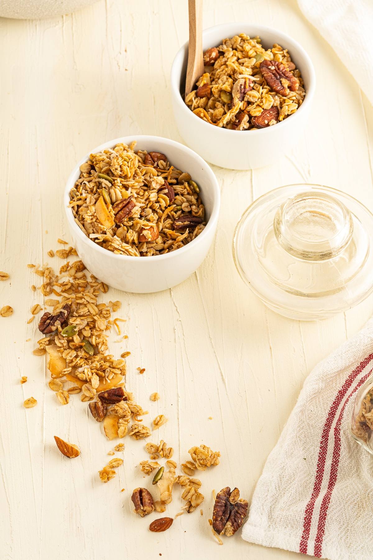 Two white bowls of easy homemade granola with spilled granola on a light surface, next to a glass jar and a white towel—part of a healthy breakfast.