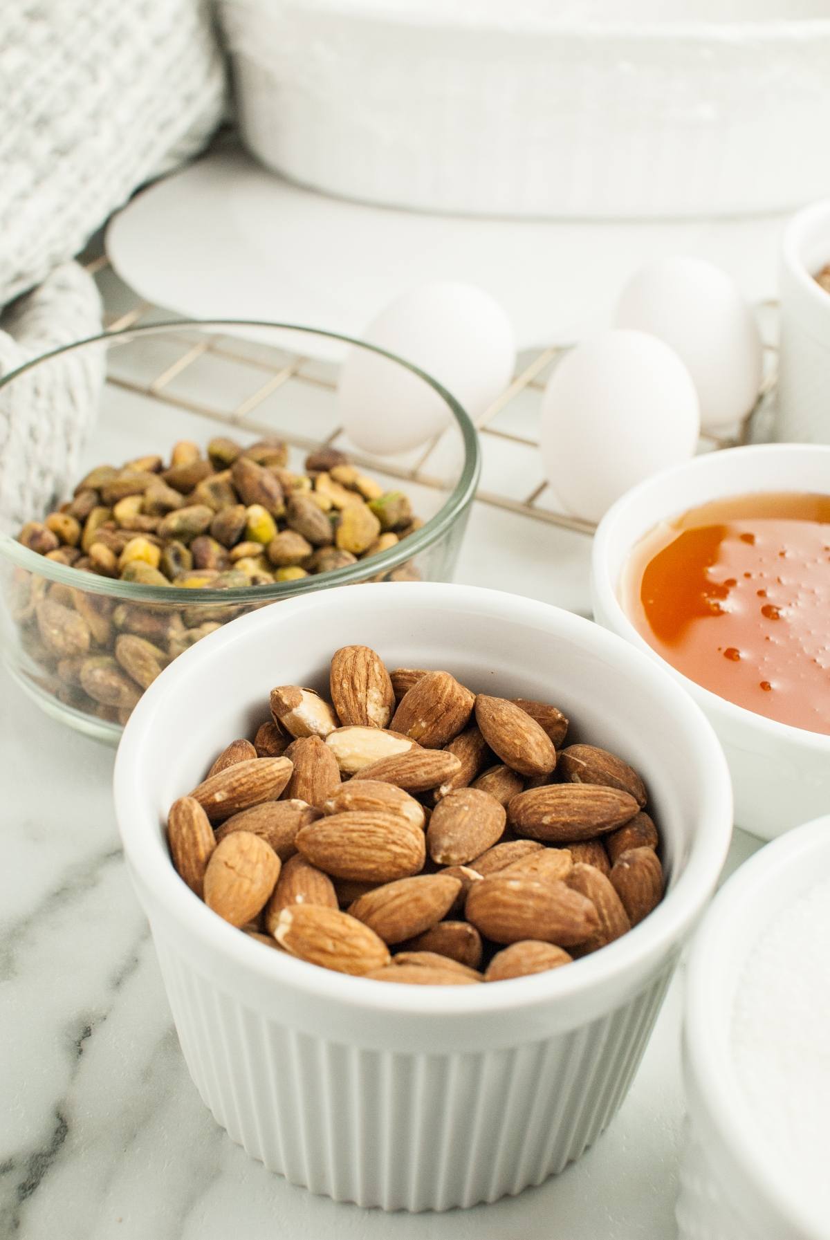 A bowl of almonds, a bowl of pistachios, eggs, and a dish of honey on a marble countertop—classic ingredients for Italian Nut Nougat, the perfect Christmas candy with roasted nuts.