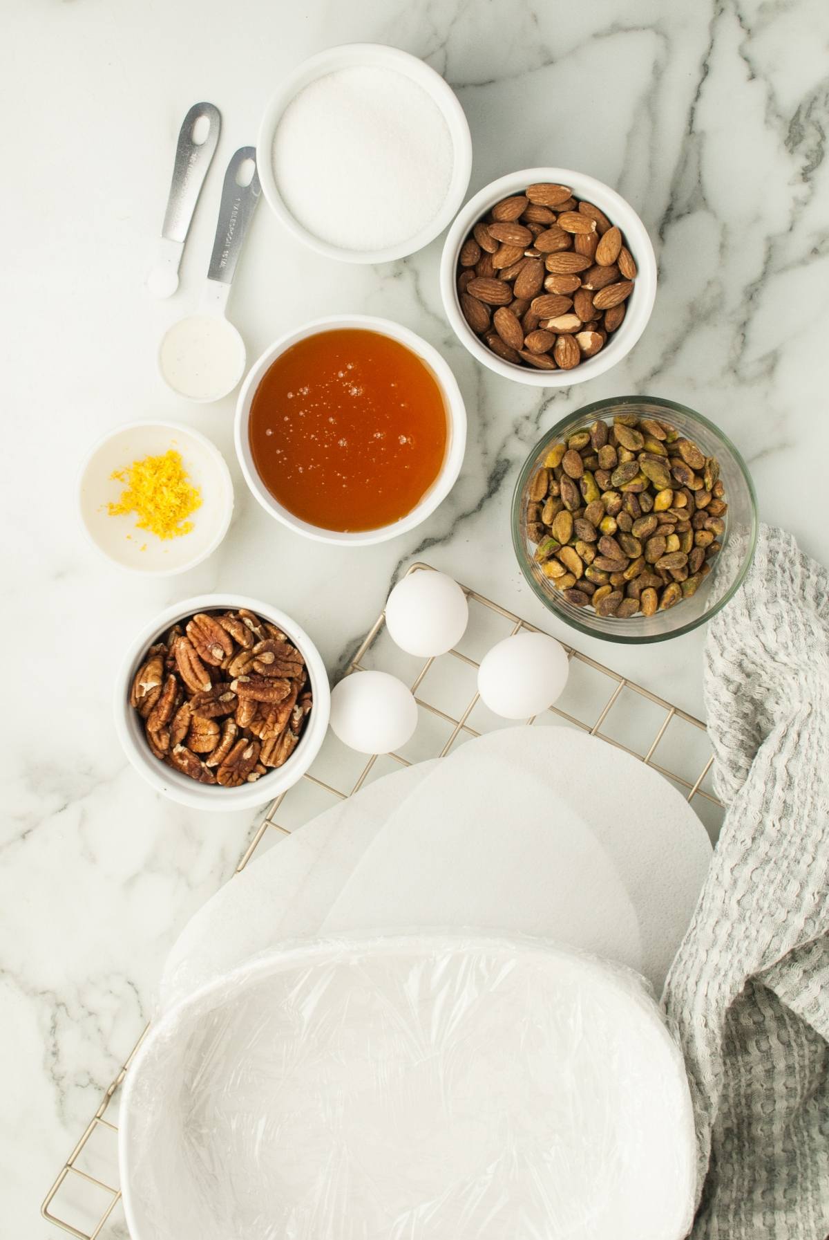 Bowls of sugar, honey, almonds, pecans, pistachios, lemon zest, eggs, and measuring spoons on a marble counter—ready to create classic Italian Nut Nougat, a favorite Christmas candy with roasted nuts.