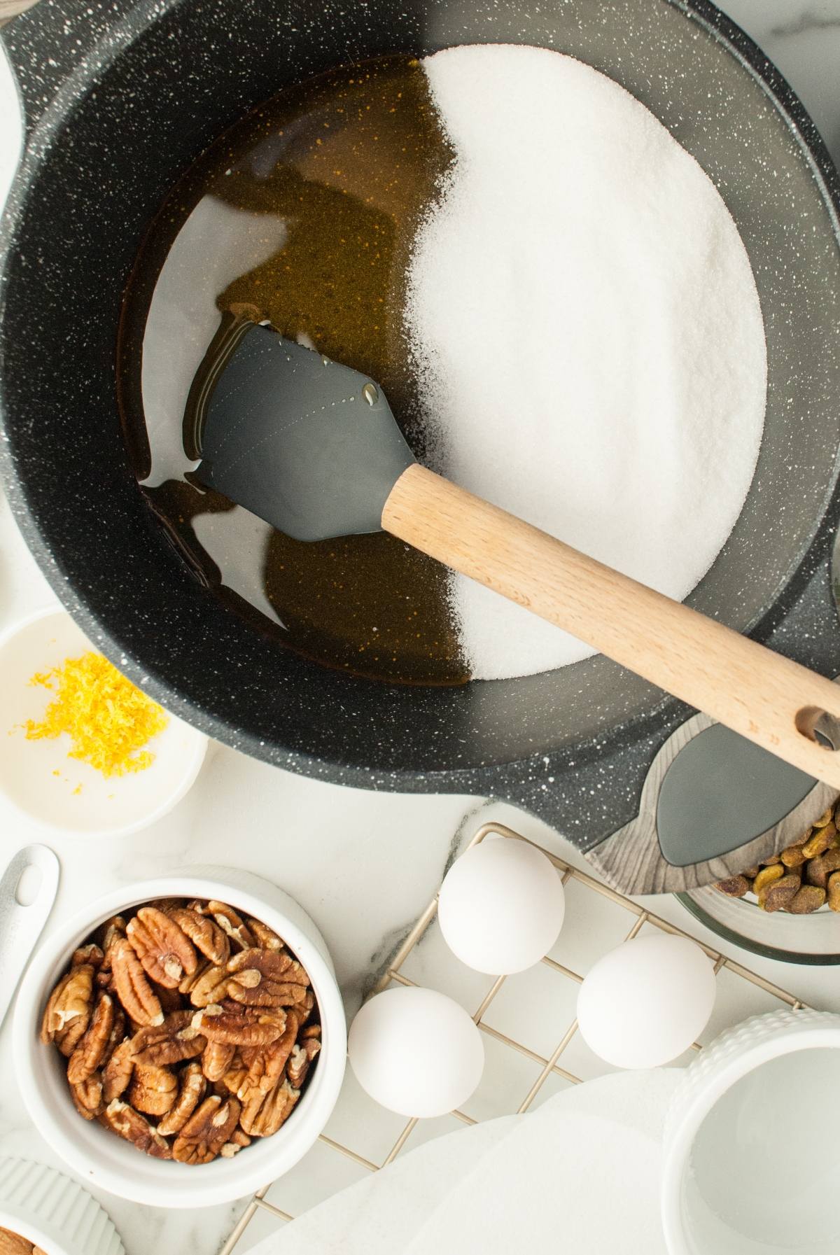 A saucepan with sugar and syrup, a spatula, eggs, pecans, zest in bowls on a white counter—perfect for making homemade Torrone Italian Nut Nougat or your favorite Christmas candy with roasted nuts.