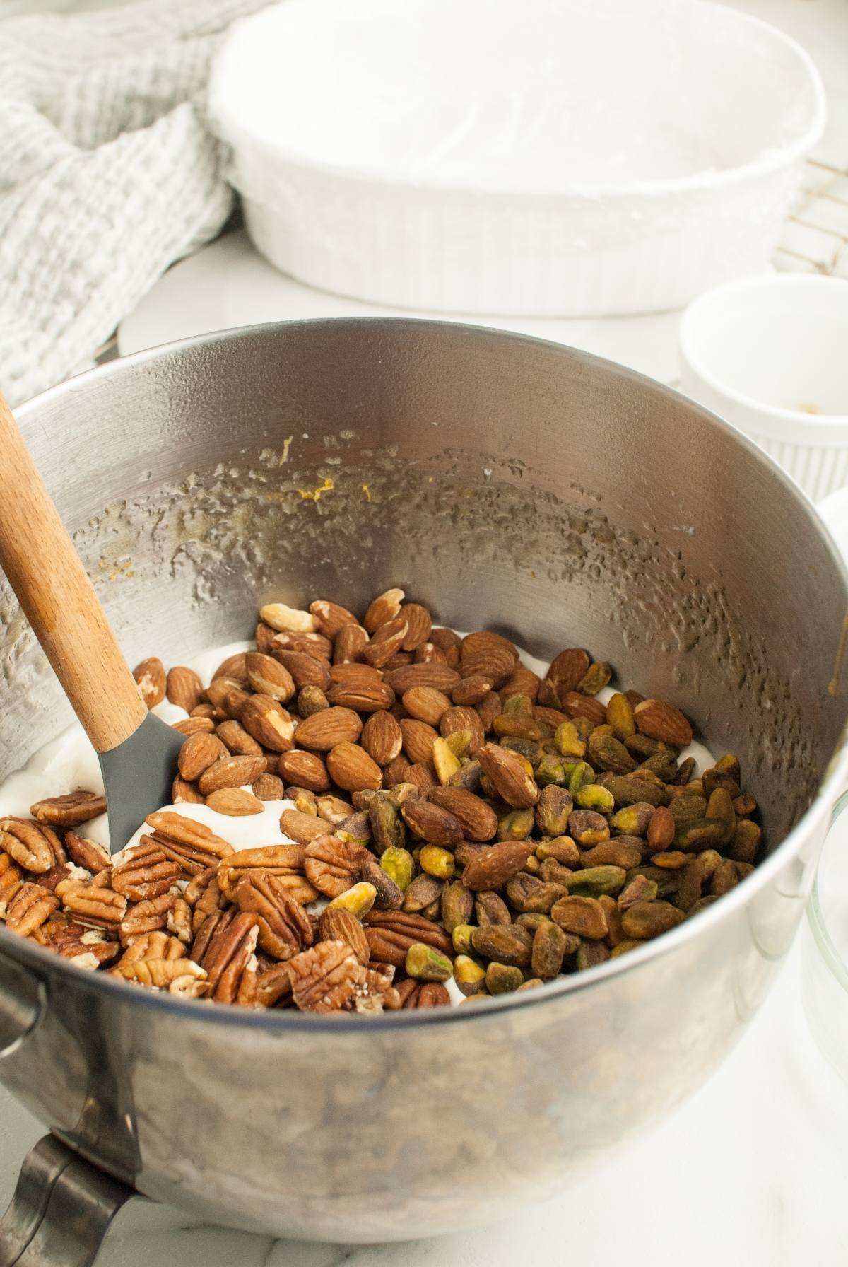 Metal mixing bowl with pecans, almonds, pistachios, and batter being mixed with a spatula on a kitchen counter while preparing Homemade Torrone Italian Nut Nougat.
