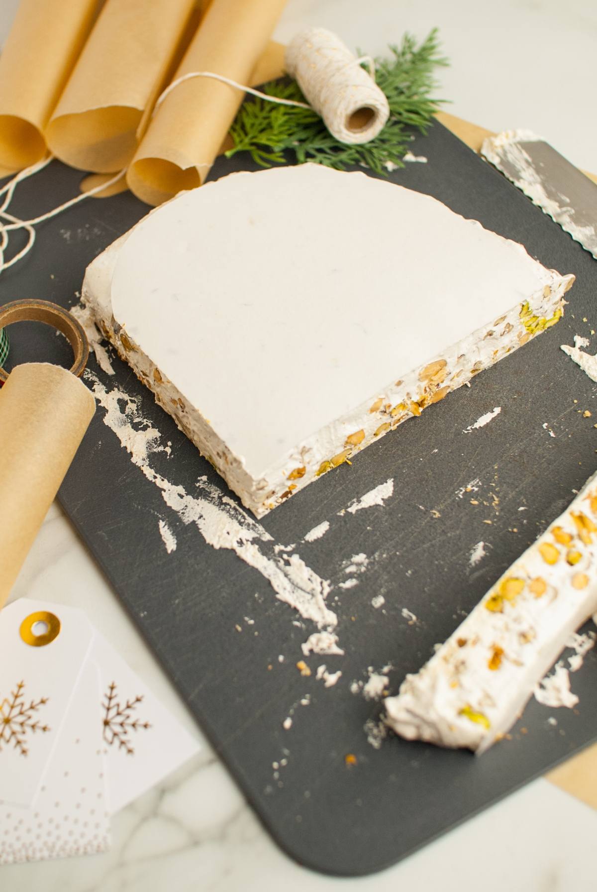 A slab of Italian Nut Nougat, the perfect Christmas candy with roasted nuts, sits on a slate board surrounded by wrapping paper, string, and gift tags.
