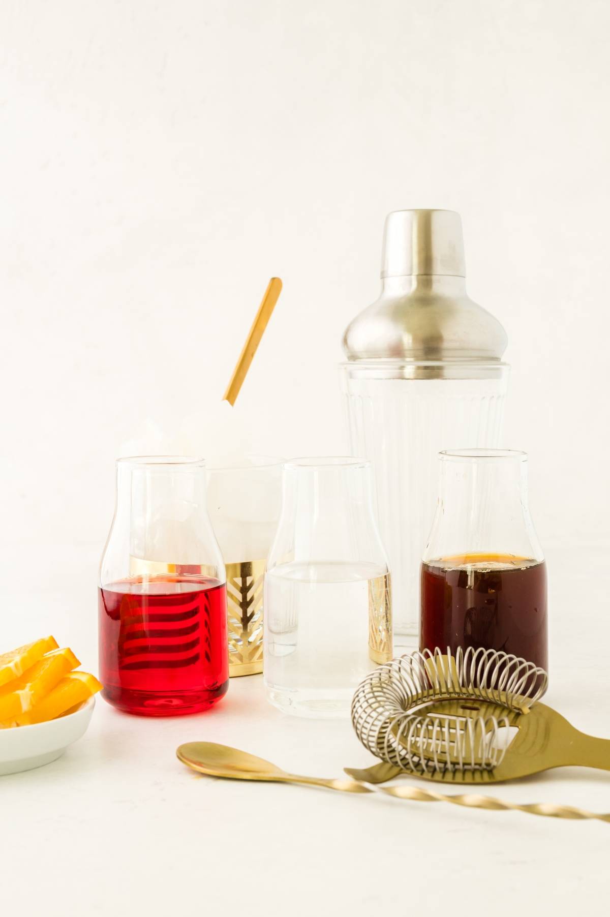 Three glass jars with red, clear, and dark liquids hint at a classic negroni cocktail. A cocktail shaker, bar tools, and fresh orange slices on a plate complete this inviting setup for crafting a perfect cocktail with Campari.