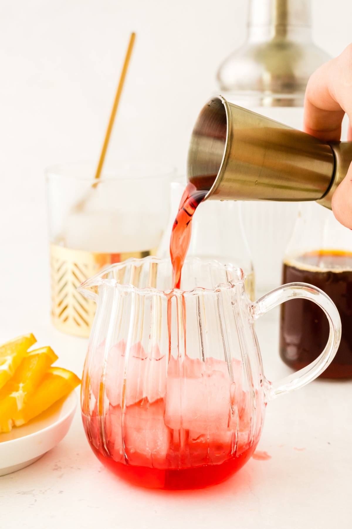 A hand pours a classic Italian cocktail—red negroni—into a glass pitcher with ice, while orange slices and drinks sit nearby.