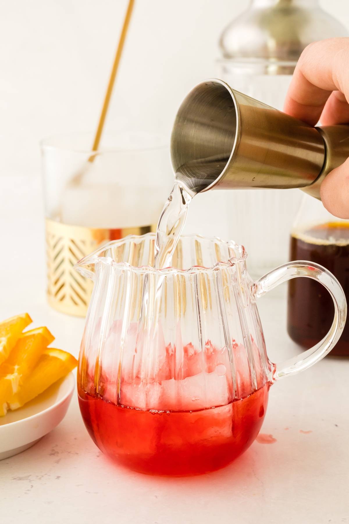 Hand pours clear liquid into a glass pitcher with ice and red drink; orange slices on the side—perfect for mixing a classic Italian cocktail like the negroni cocktail.