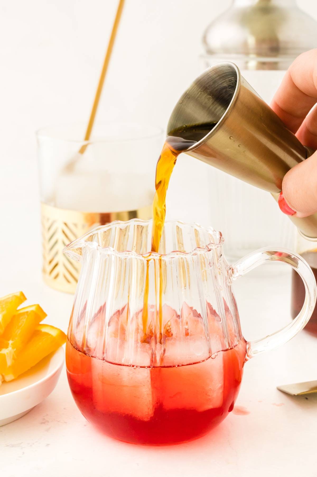 A hand pours dark liquid into a glass pitcher with red drink and ice, next to orange slices—perfect for mixing a classic Italian cocktail with Campari.