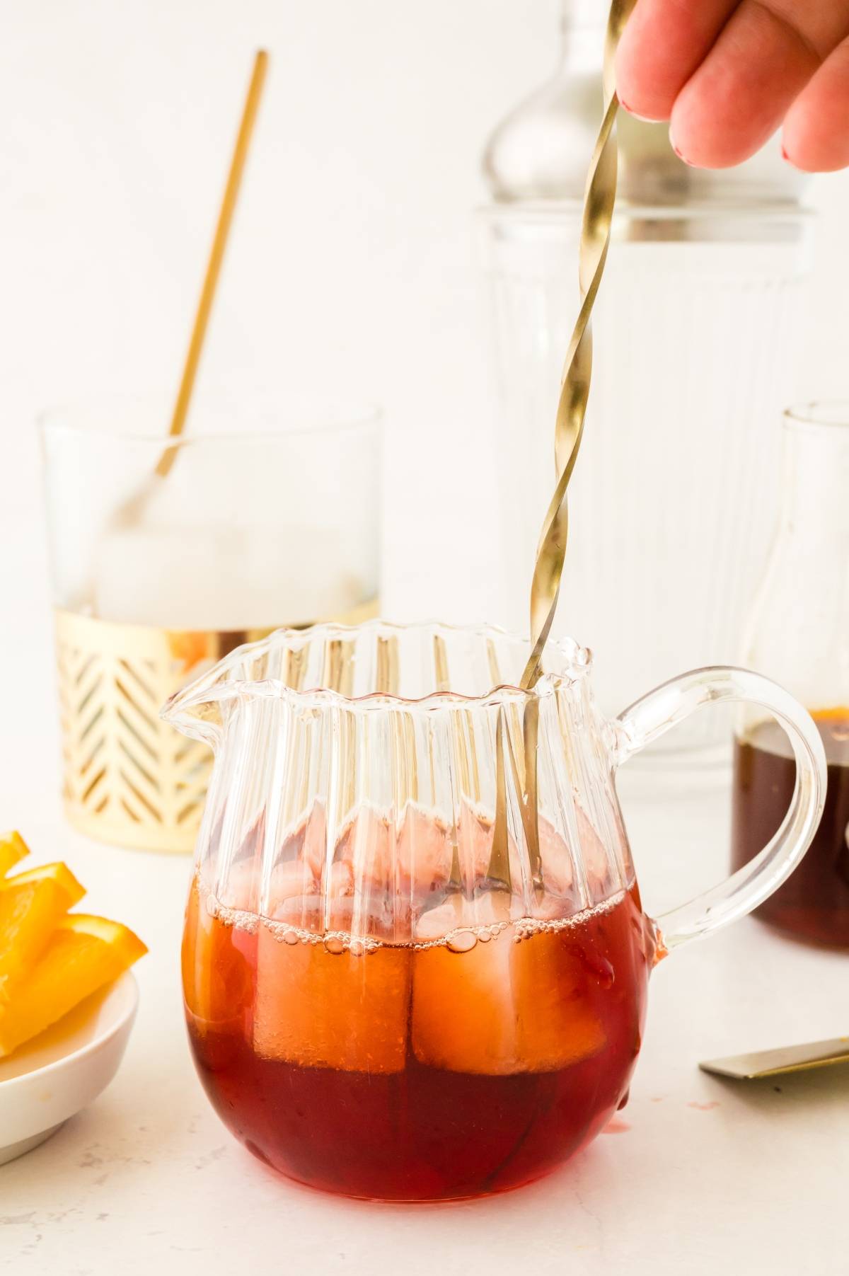A hand stirs a red negroni cocktail with ice in a glass pitcher; glassware and orange slices are nearby.