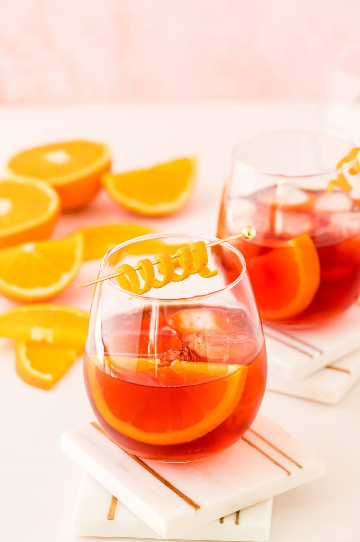 Two glasses of classic Italian cocktail with ice, orange slices, and a curled orange garnish, with orange wedges nearby—perfect for enjoying a refreshing negroni cocktail.
