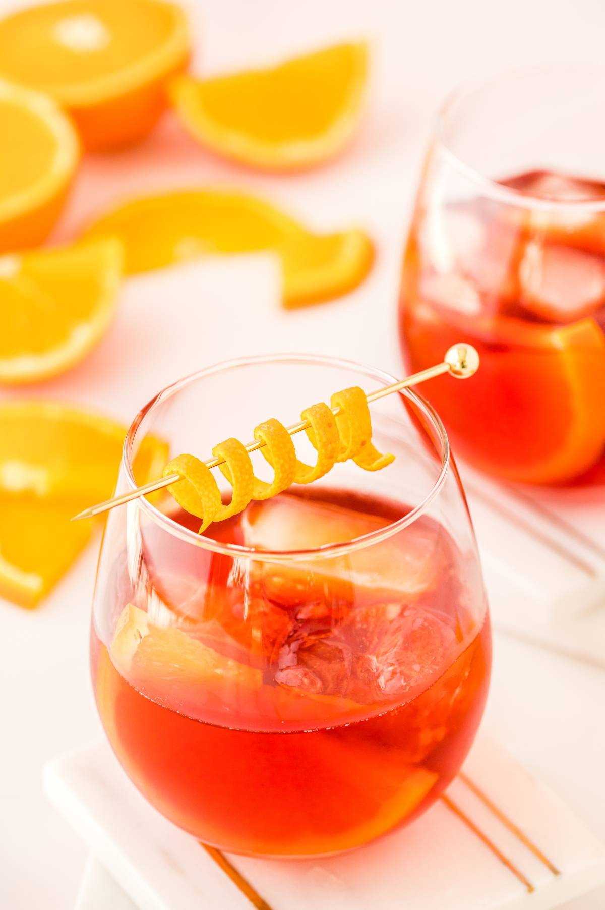 A glass of orange cocktail with ice, garnished with an orange twist, and orange slices in the background—perfect for enjoying a classic negroni cocktail or any refreshing cocktail with Campari.