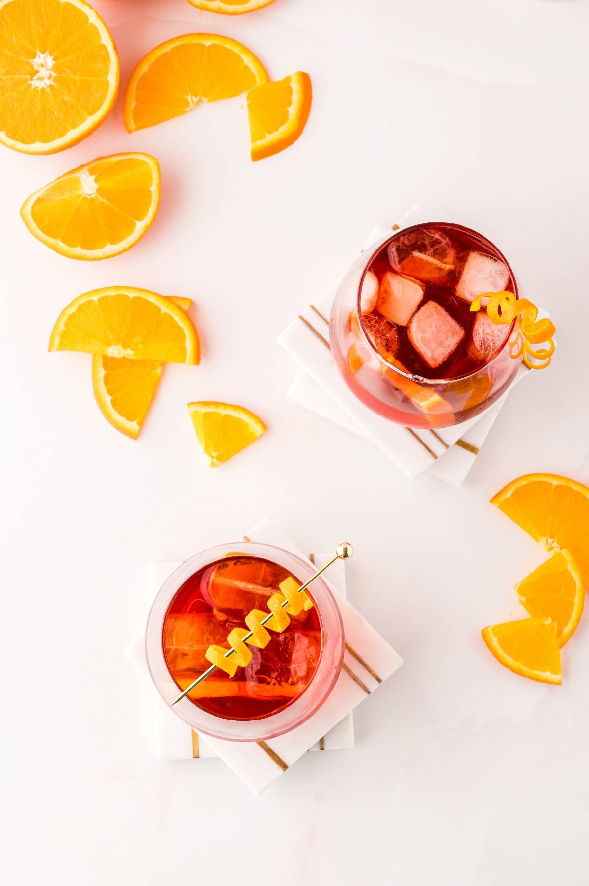 Two glasses of iced red negroni cocktails with orange garnishes, surrounded by orange slices on a white surface—a classic Italian cocktail with Campari.