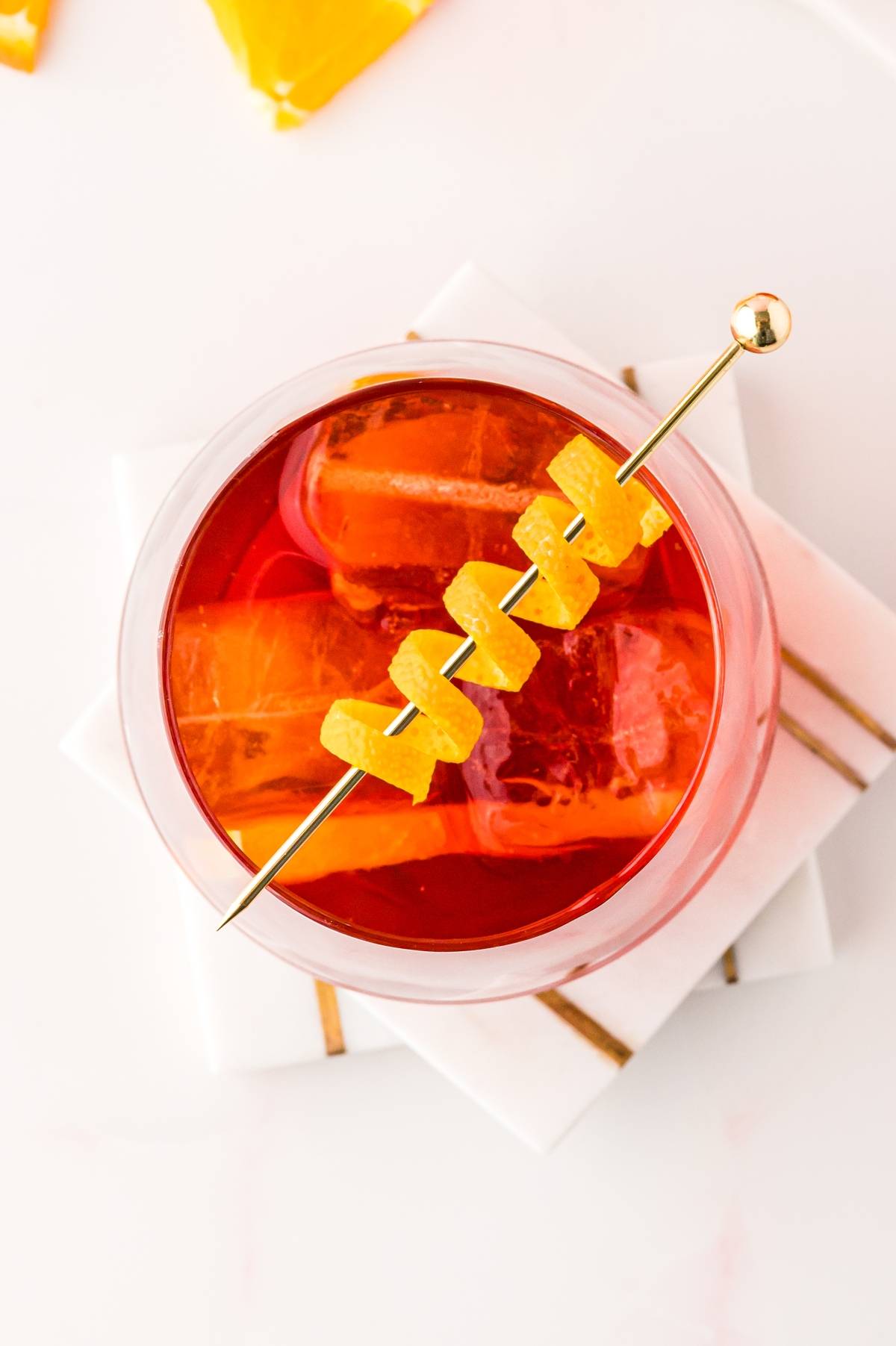 Top view of a Negroni cocktail—a classic Italian cocktail with Campari—served over ice and garnished with orange peel on a skewer, resting on white coasters.