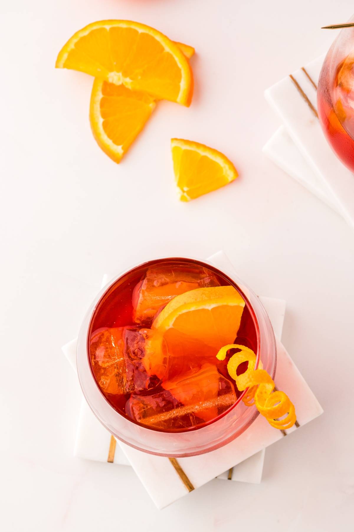 Top view of a negroni cocktail with ice and an orange slice, garnished with a citrus twist, on white coasters—perfect inspiration for learning how to make the best negroni at home.