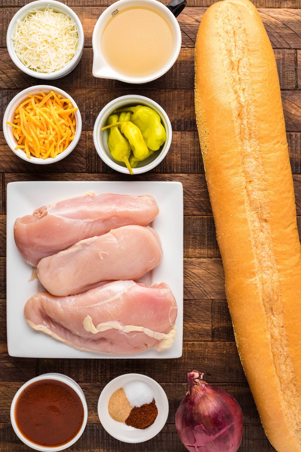 Ingredients for a sandwich: raw chicken, baguette, cheeses, peppers, onion, broth, spices, and sauce on a wooden board—perfect for making an easy chicken pizza or a tasty barbeque French bread pizza.