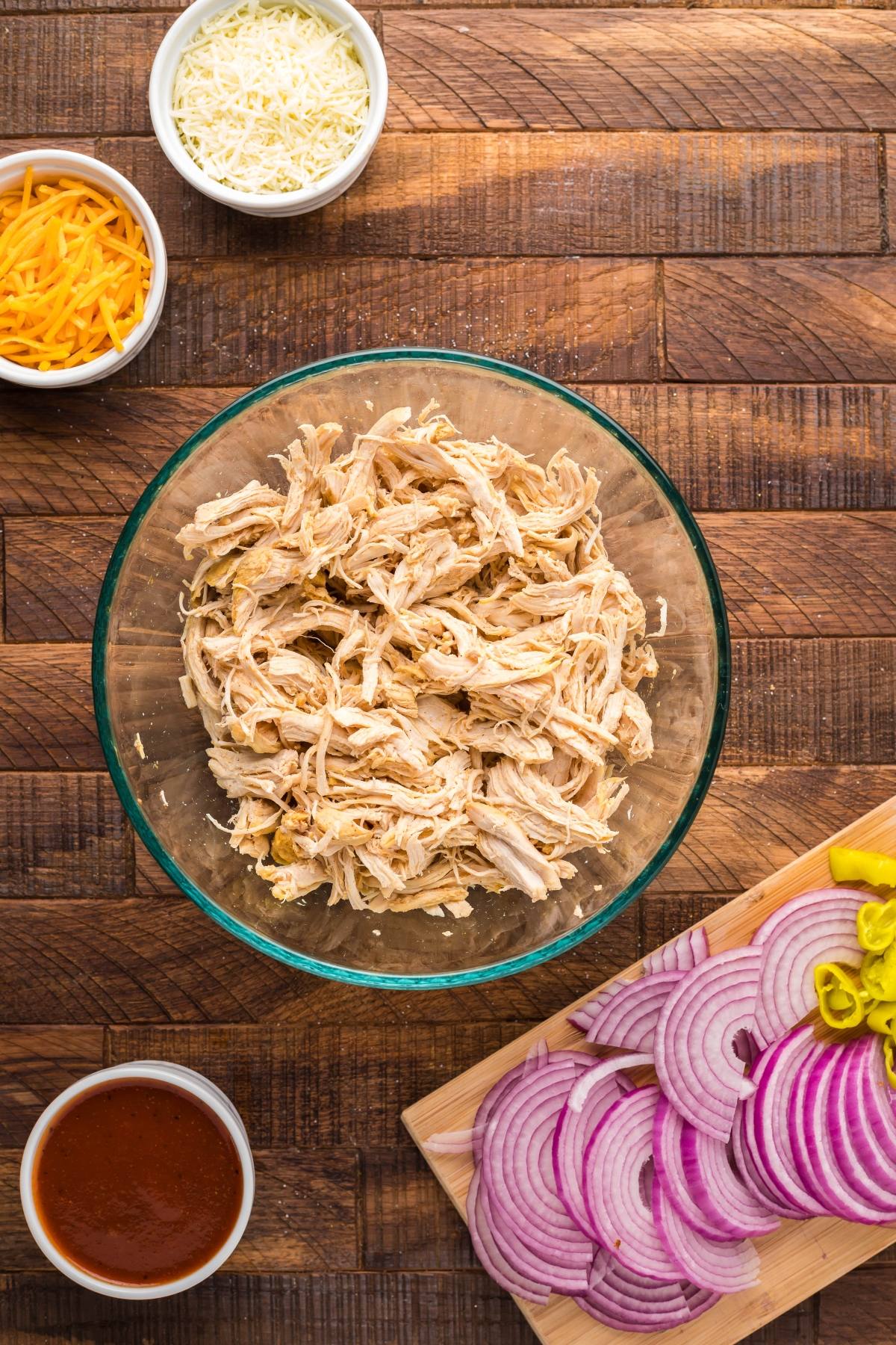 A bowl of shredded chicken, sliced red onions, cheese, sauce, and peppers arranged on a wooden table—perfect toppings for an easy chicken pizza or a flavourful Instant Pot dinner.