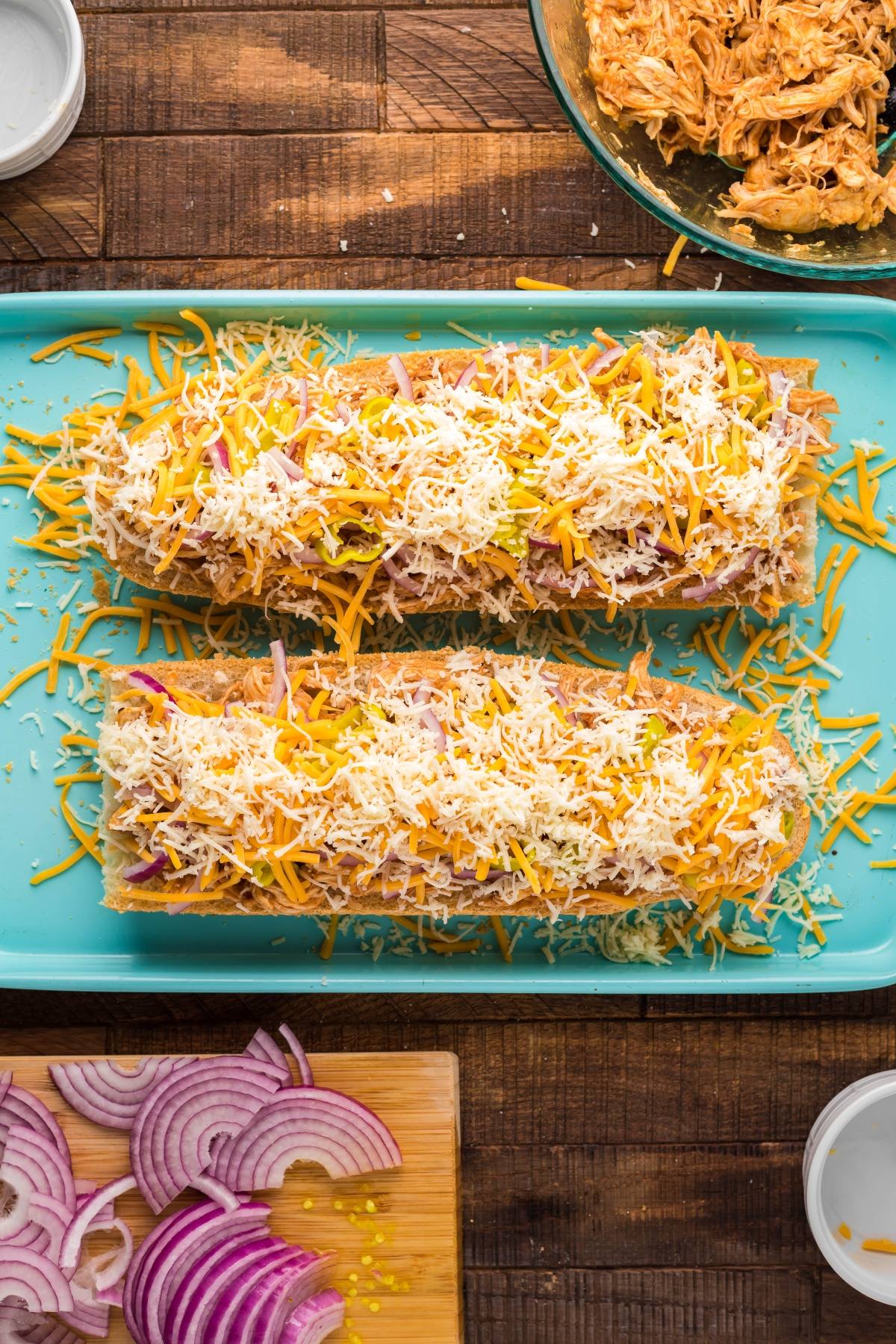 Two stuffed baguettes topped with shredded cheese on a blue tray, with onions and shredded chicken nearby—an Easy chicken pizza twist that makes for a perfect 20 minute meal.
