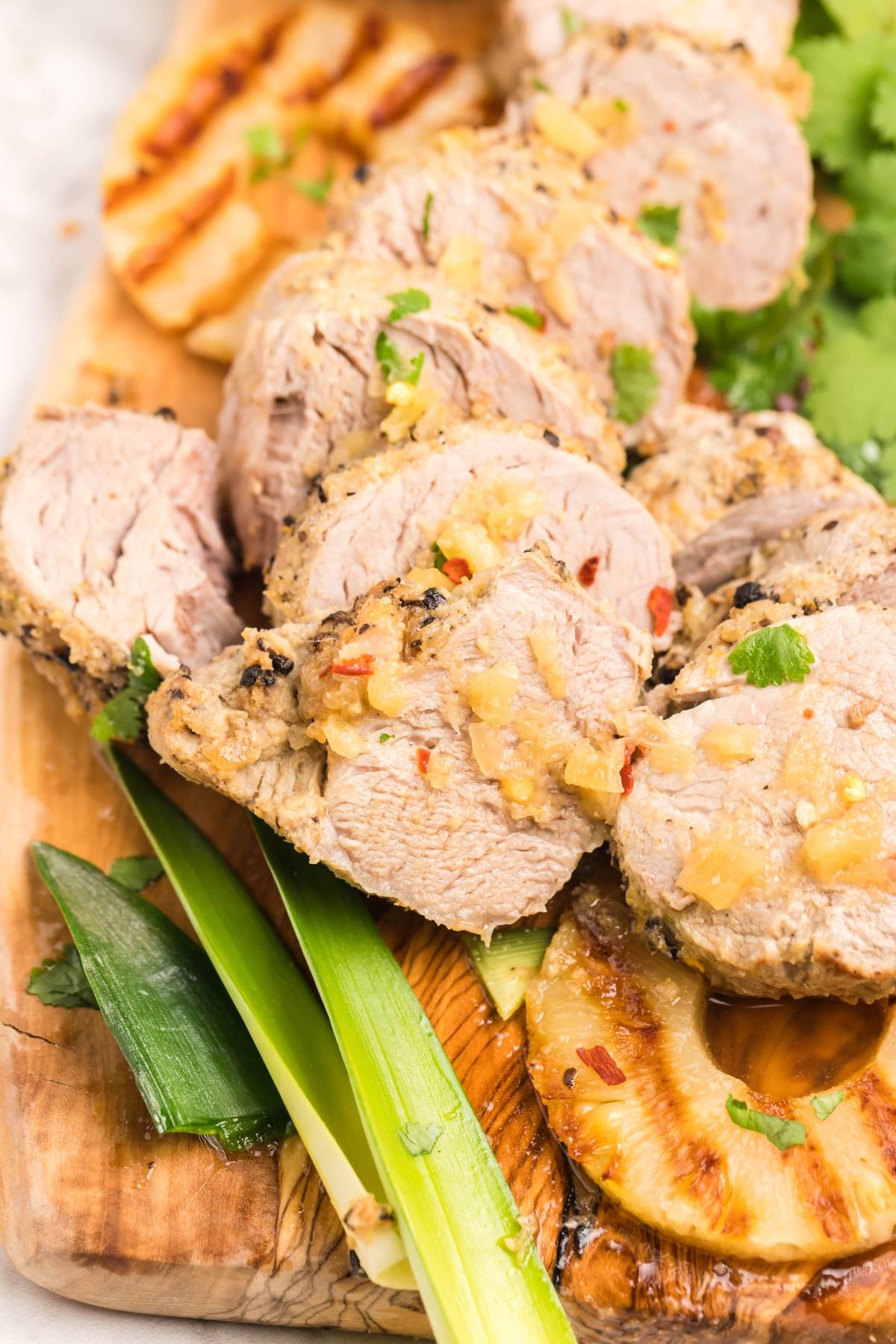 Sliced roasted Instant Pot pork tenderloin with herbs on a wooden board, garnished with grilled pineapple and green onions for a Hawaiian inspired recipe.
