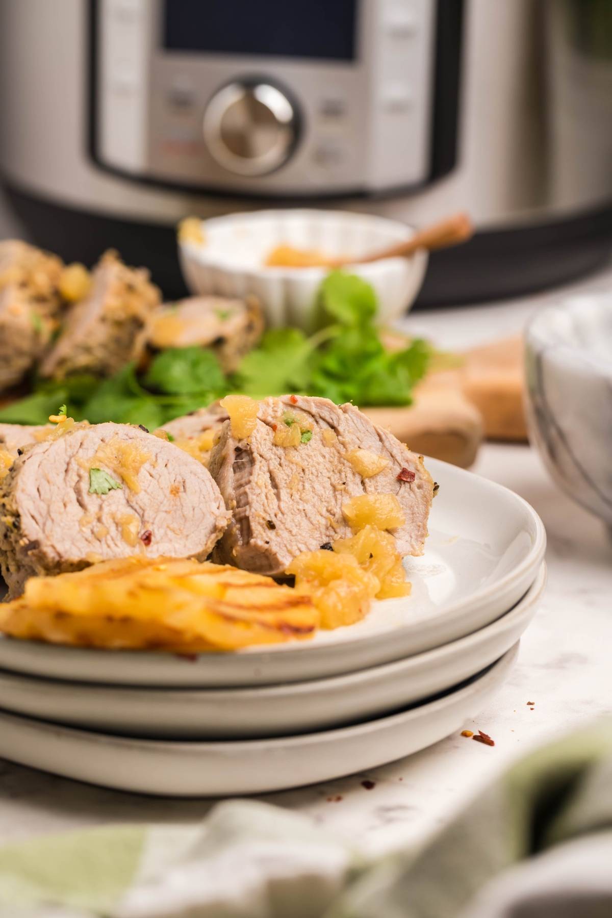 Sliced Instant Pot Hawaiian pork tenderloin with pineapple on white plates, with an Instant Pot and herbs in the background.