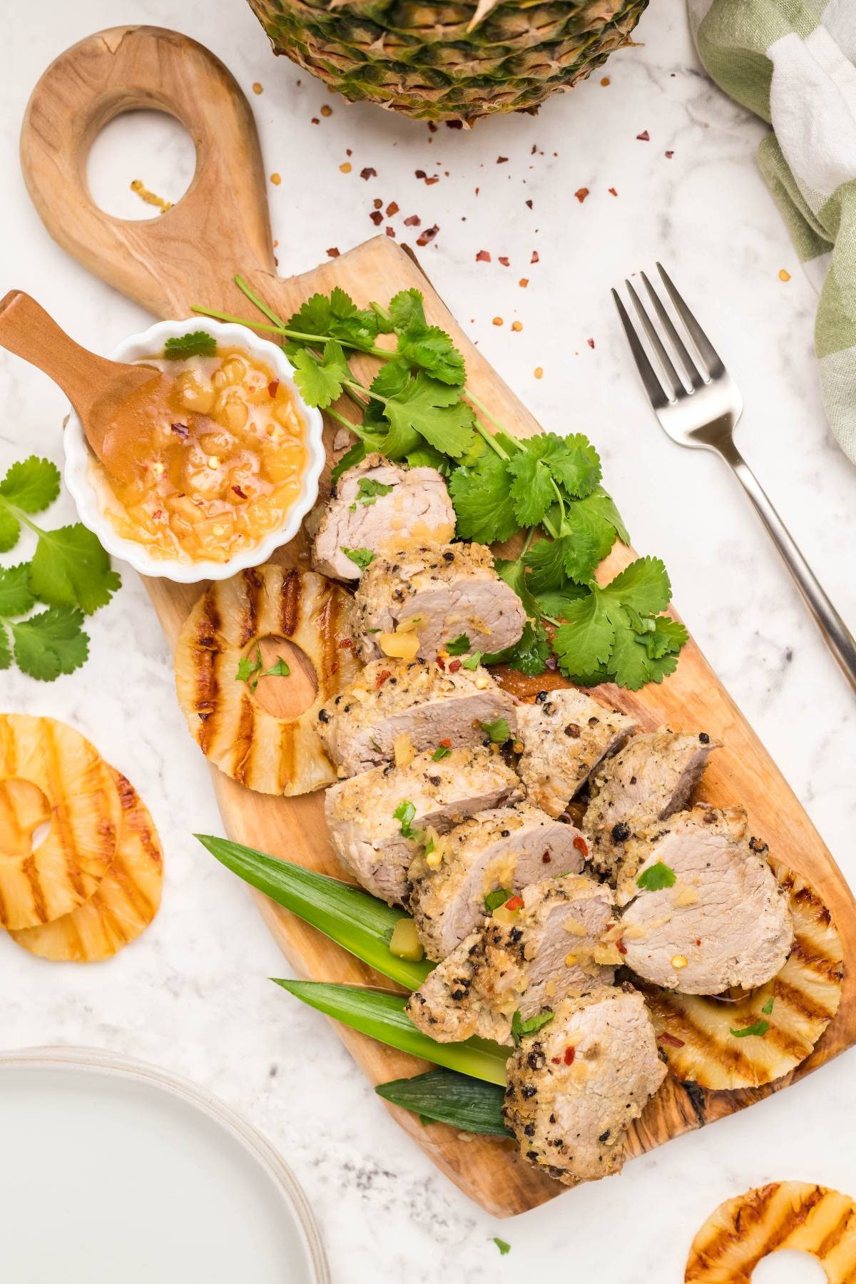 Sliced grilled pork tenderloin on a wooden board with grilled pineapple, cilantro, and dipping sauce—an easy Hawaiian inspired recipe perfect for any dinner.