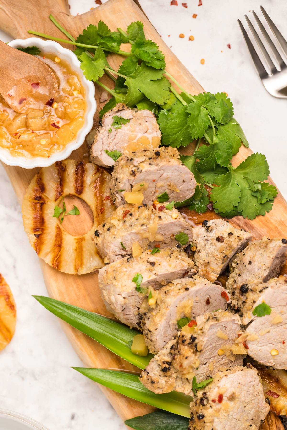 Sliced pork tenderloin with grilled pineapple, fresh cilantro, and sauce on a wooden board—an easy Hawaiian inspired recipe bursting with tropical flavors.