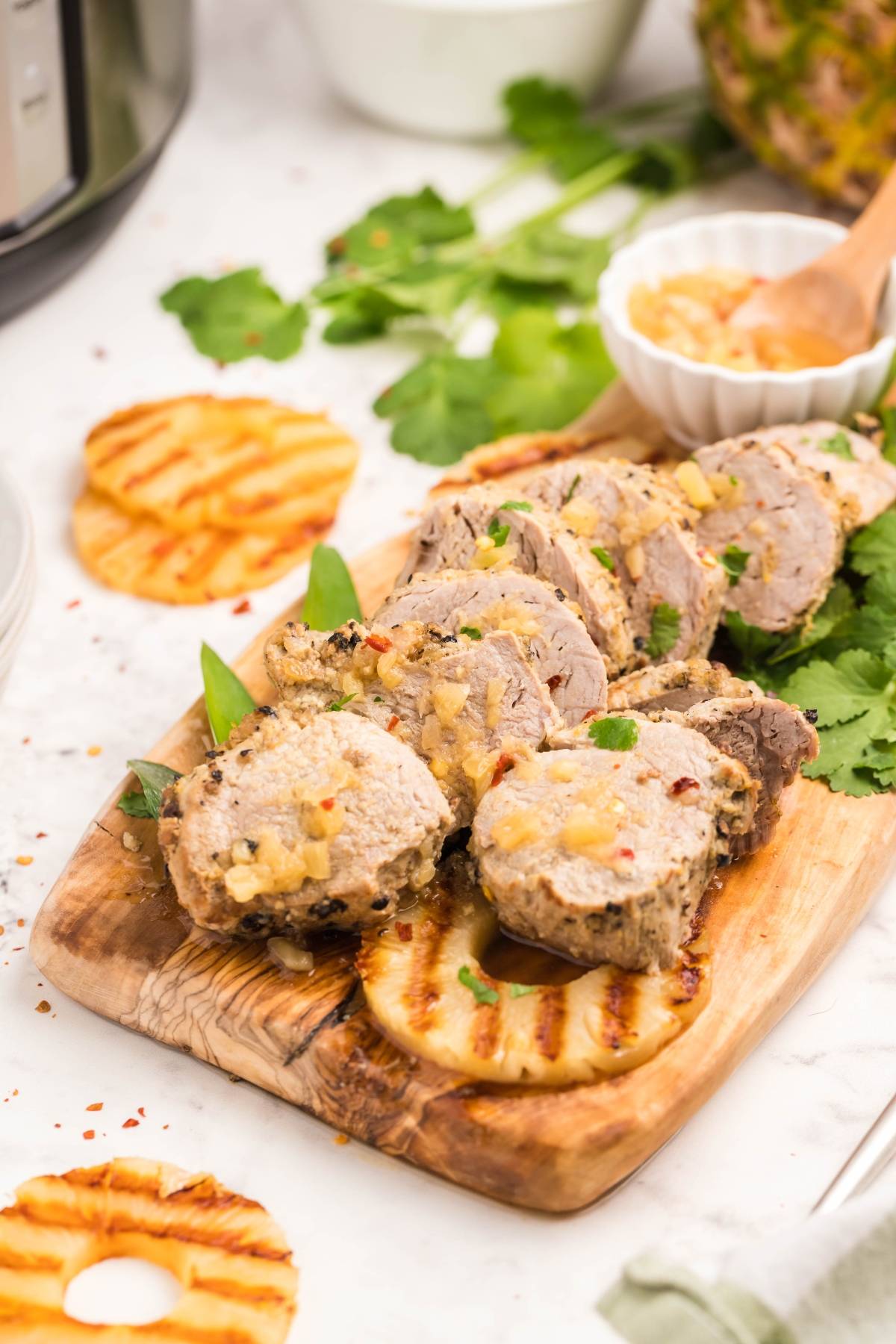 Sliced Instant Pot Hawaiian pork tenderloin on a wooden board with grilled pineapple and fresh herbs, served on a white table—an easy instant pot dinner recipe with delicious Hawaiian inspired flavors.