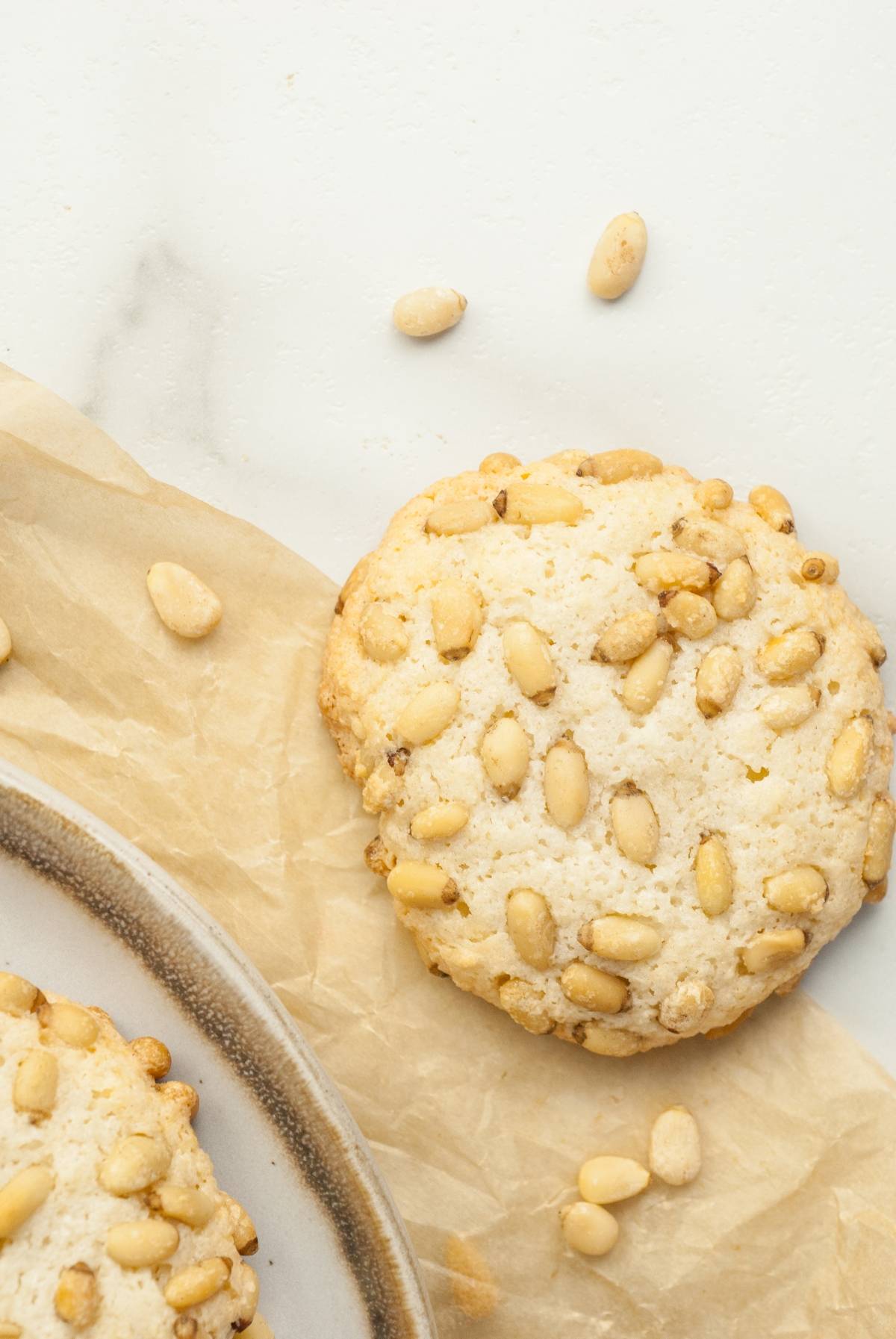 A pine nut cookie on parchment paper with scattered pine nuts on a white surface, showcasing classic Italian Pignoli Cookies—beloved almond and pine nut cookies perfect for any sweet occasion.