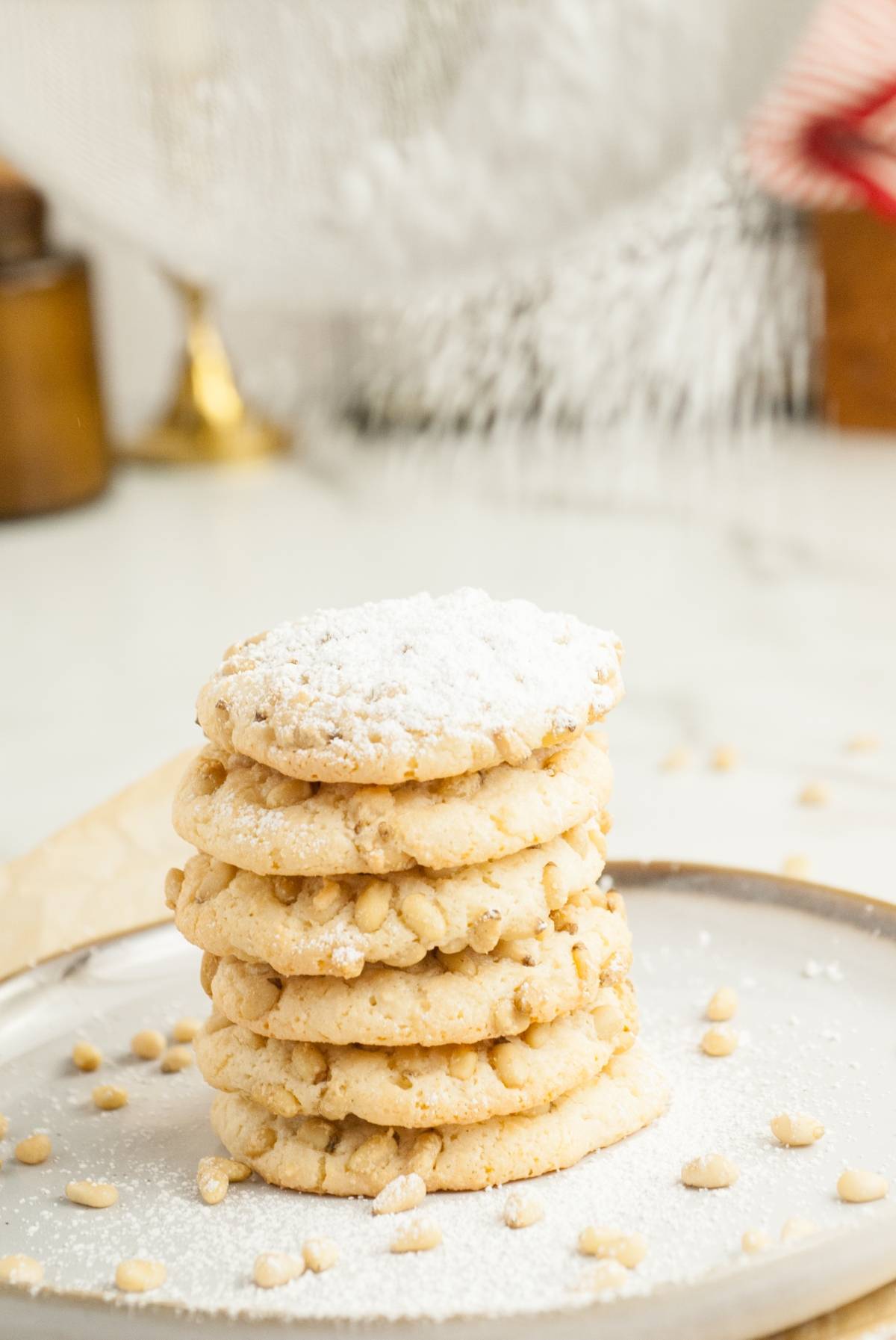 A stack of Italian Pignoli Cookies dusted with powdered sugar sits on a plate, surrounded by scattered pine nuts.