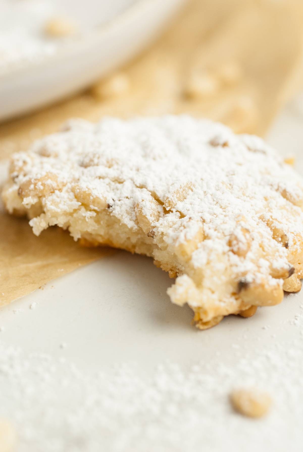 A powdered sugar cookie with a bite taken out, resting on parchment paper—perfect for easy gluten free holiday baking and reminiscent of classic almond and pine nut cookies.