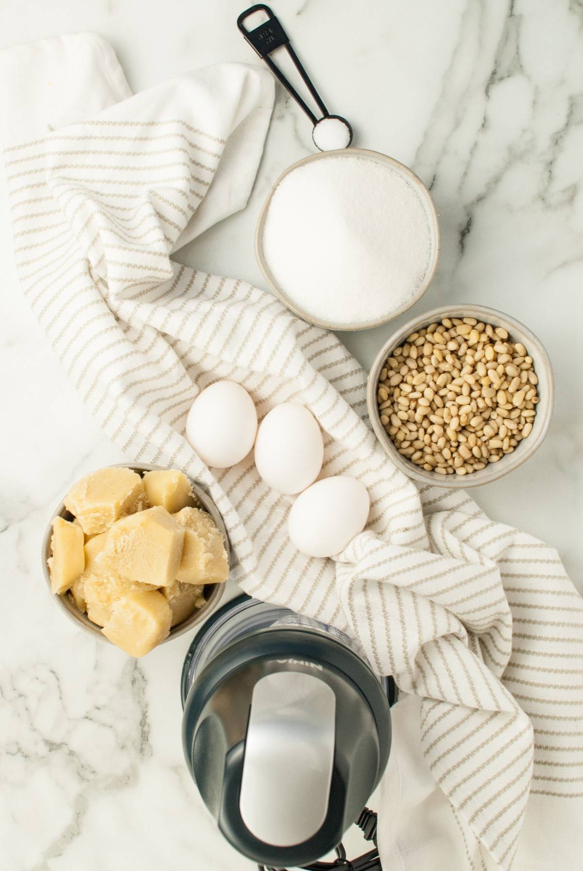 Overhead view of eggs, sugar, pine nuts, cheese, a striped towel, and a mixer on marble—perfect for making the best Italian Christmas cookies or trying an easy gluten free holiday baking recipe like classic pignoli cookies.