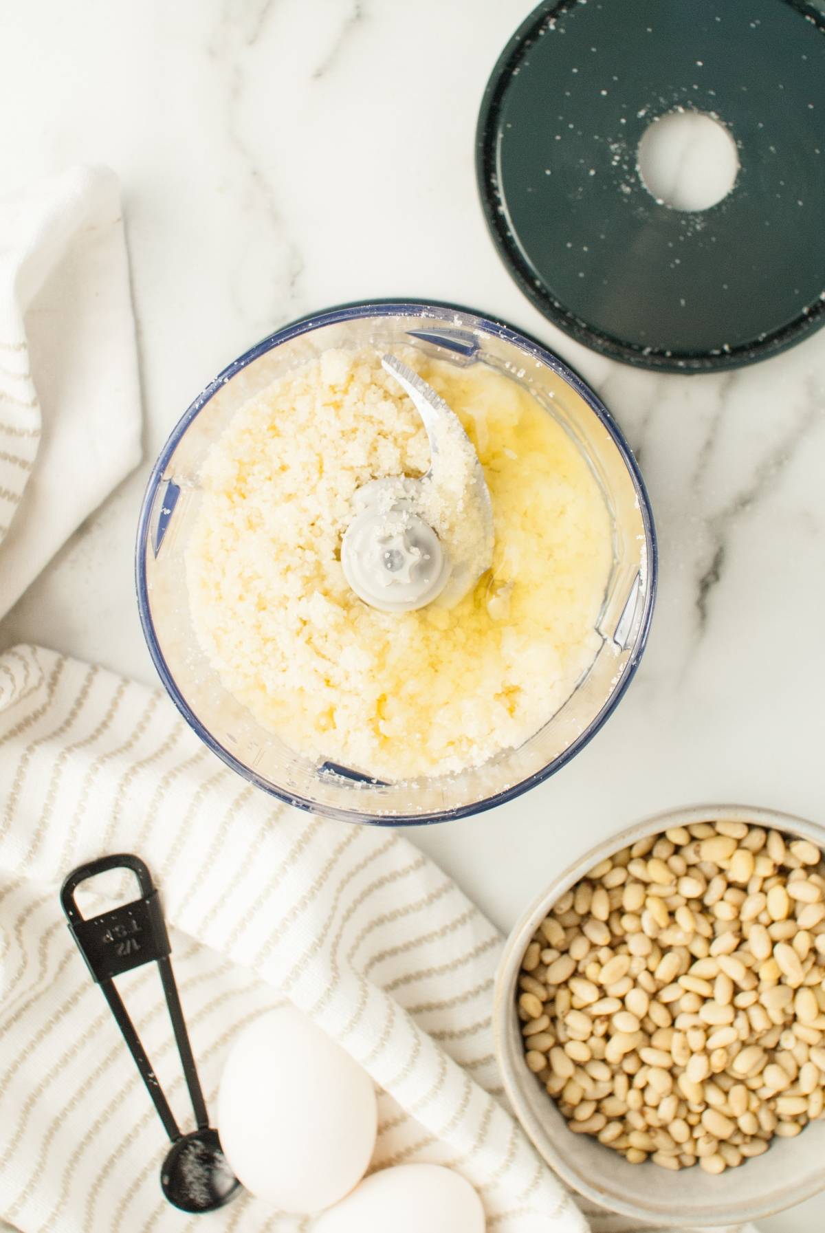 Food processor with blended ingredients, eggs, pine nuts, and a measuring spoon on a marble countertop—perfect for making the best Italian Christmas cookies or easy gluten free holiday baking like almond and pine nut cookies.