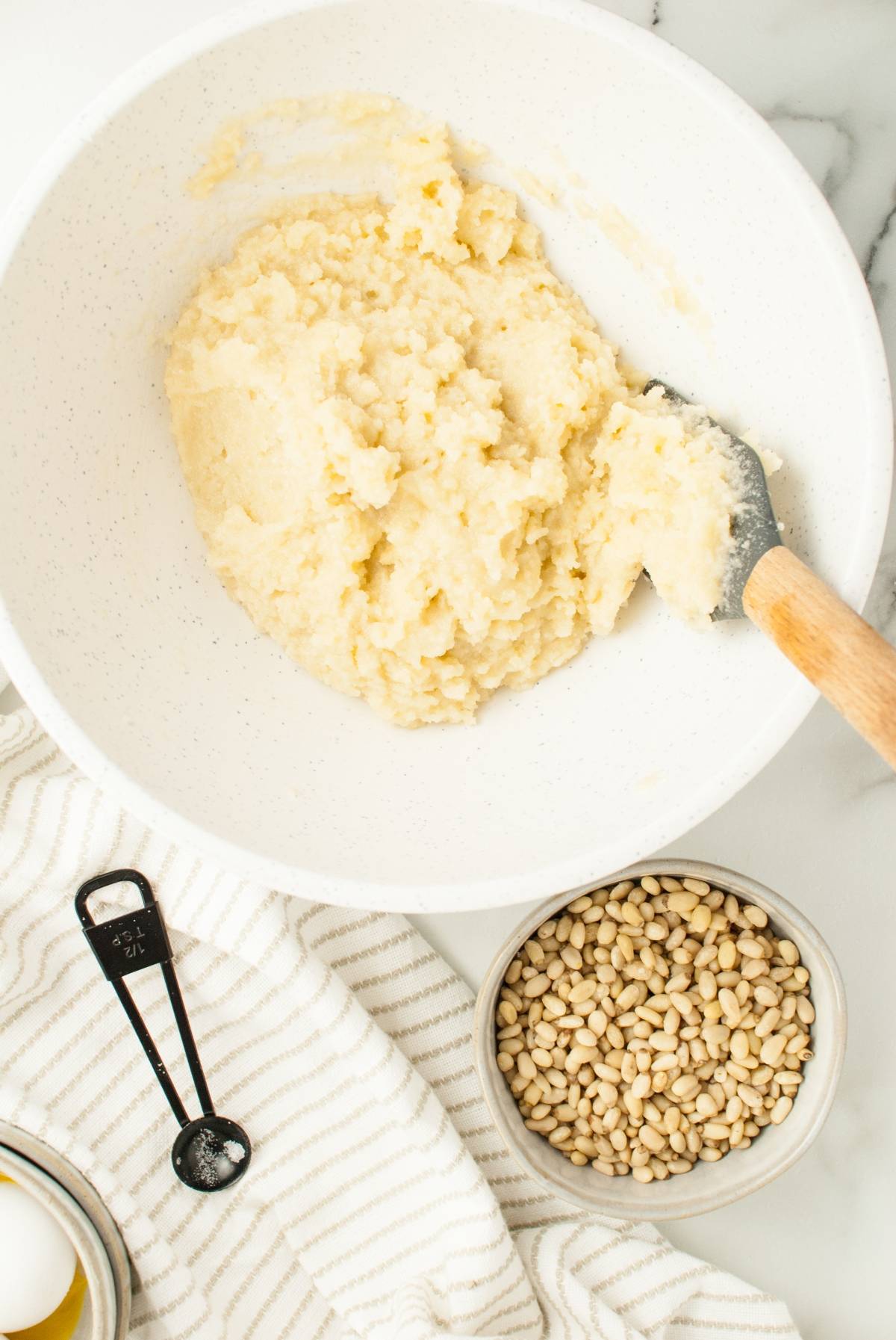 A bowl of cookie dough with a spatula, next to pine nuts, an egg, and a measuring spoon on a striped cloth—perfect for making classic Italian Pignoli Cookies or other almond and pine nut cookies.