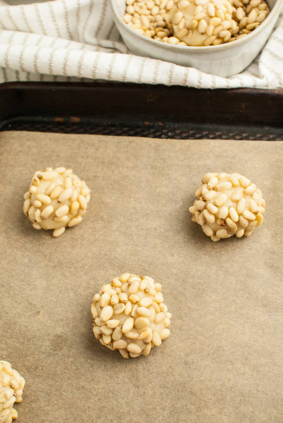 Round dough balls coated in pine nuts on a parchment-lined baking sheet, with a bowl of pine nuts in the background—classic Italian Pignoli Cookies ready to bake.