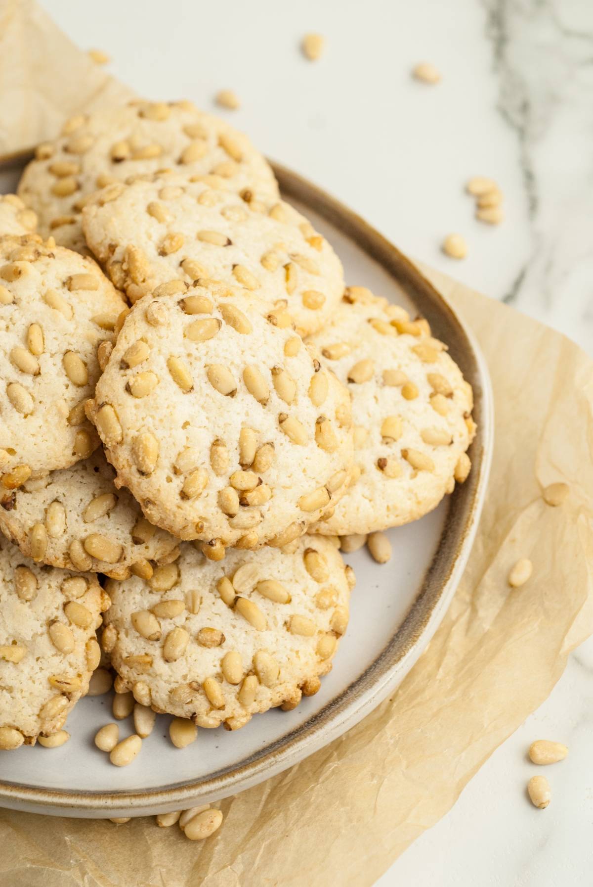 A plate of round Italian Pignoli Cookies topped with pine nuts sits on parchment paper, with extra pine nuts scattered around.