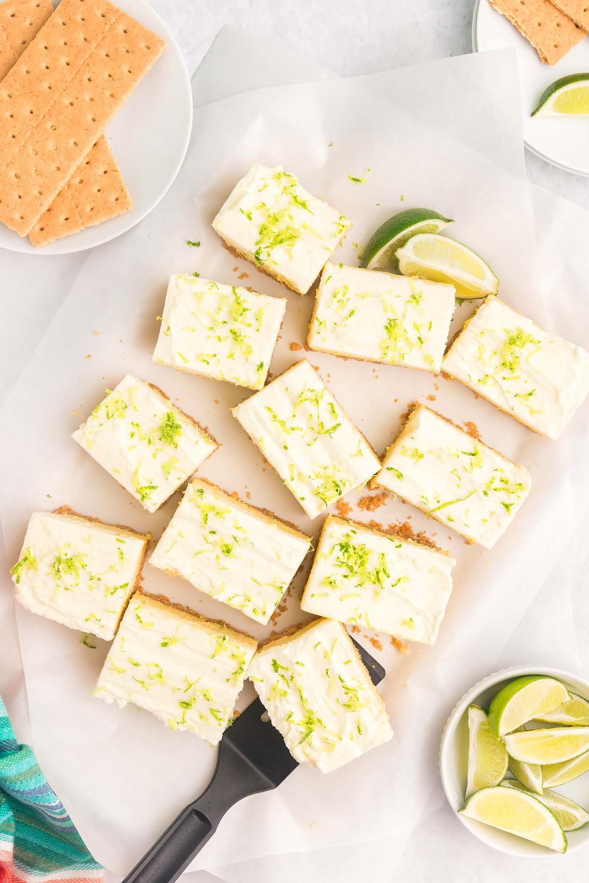 Twelve frosted key lime bars on parchment paper with lime wedges and graham crackers on the side.
