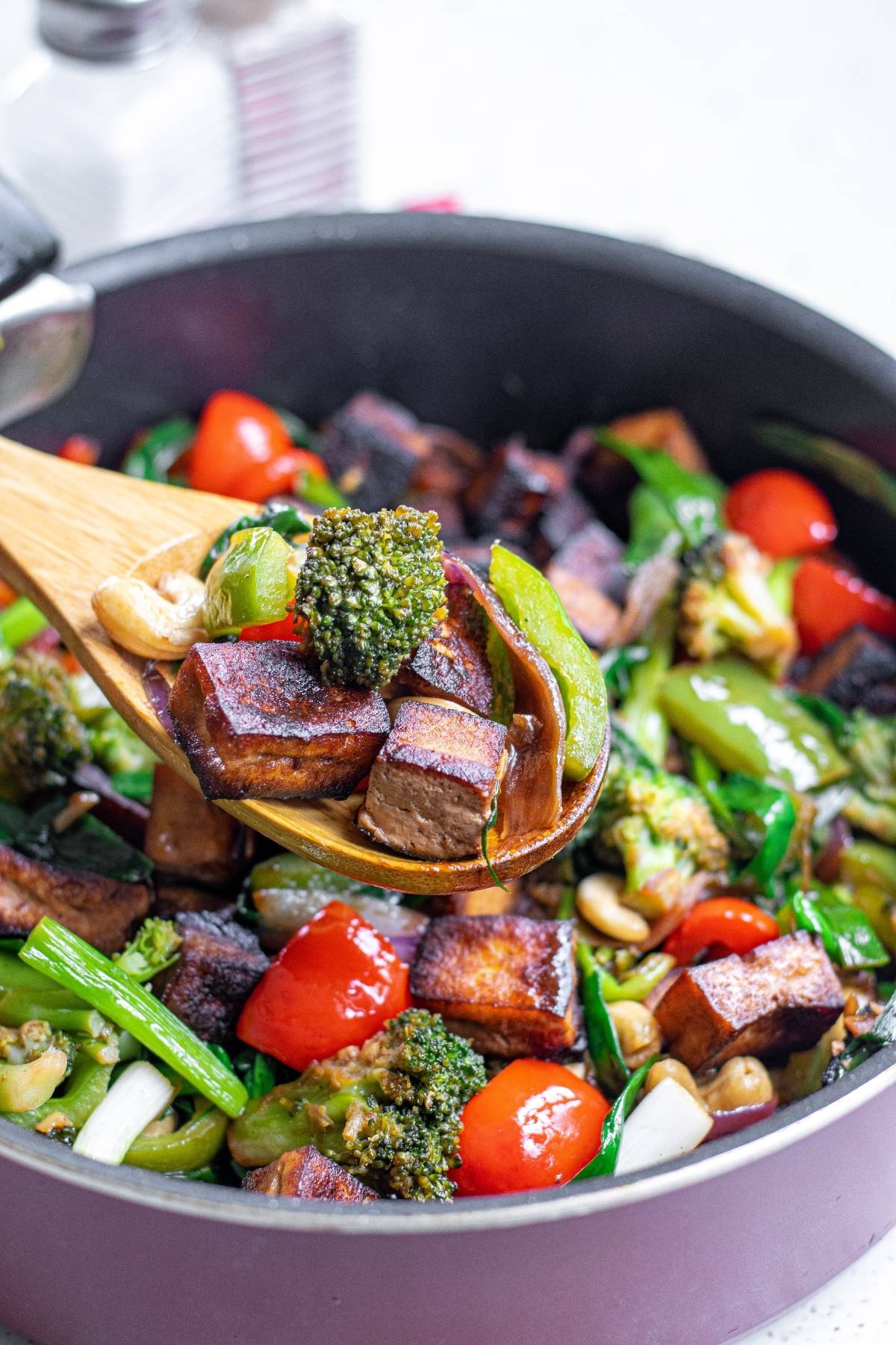 A wooden spoon holds tofu, broccoli, and vegetables over a skillet of colorful stir-fry—perfect for a vegan Kung Pao or any Chinese food tofu recipe.