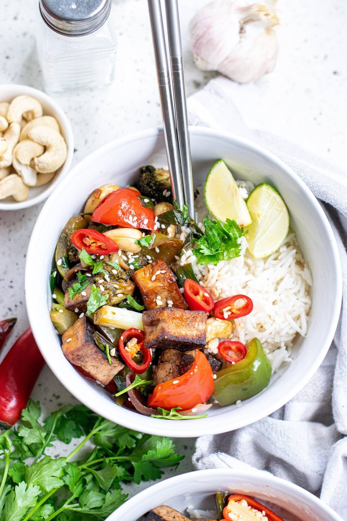 Bowl of rice with stir-fried tofu, bell peppers, broccoli, chili, lime wedges, and cilantro with chopsticks—a delicious Vegan Kung Pao perfect for a Vegetarian Chinese inspired dinner.