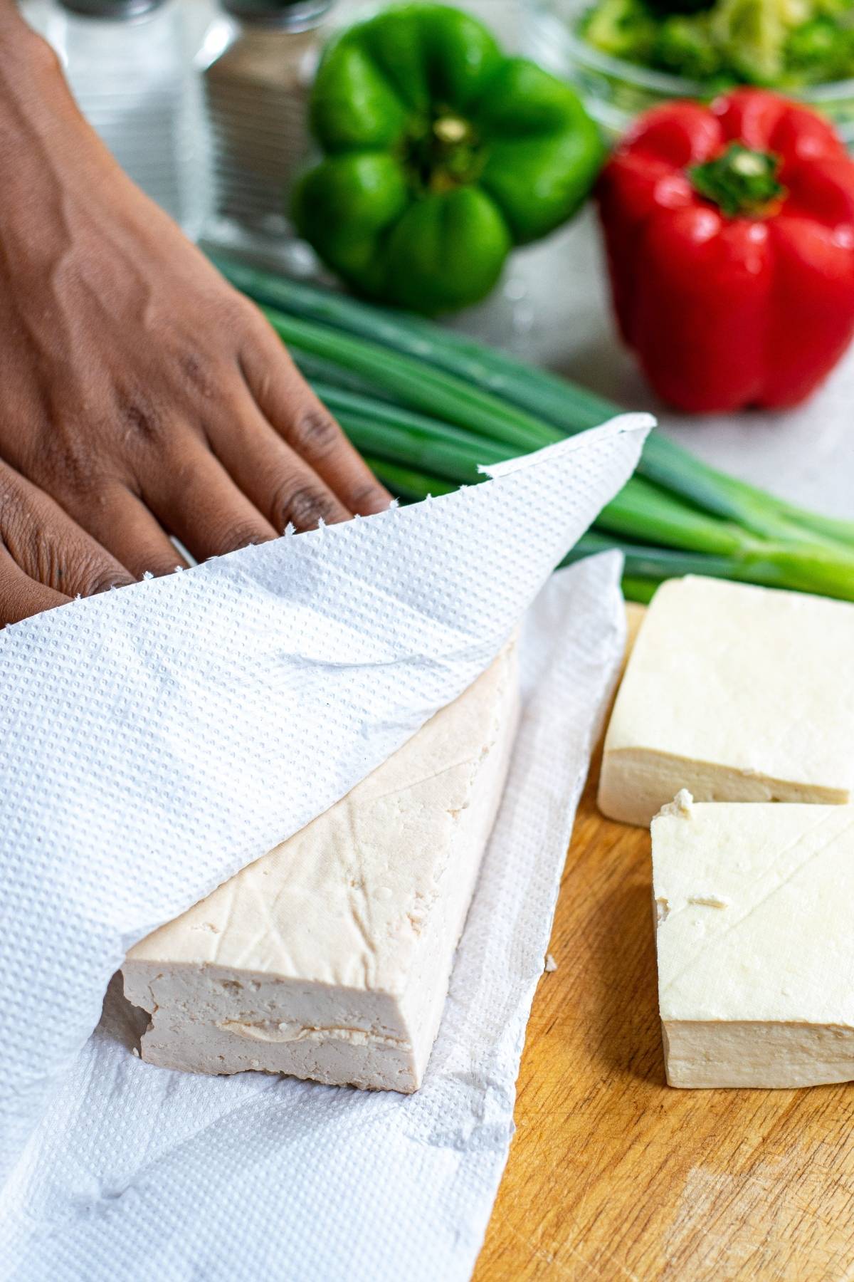 A hand presses a tofu block in a paper towel with vegetables on the table in the background, prepping for a delicious Vegan Kung Pao—a classic Chinese food tofu recipe perfect for your next vegan dinner.