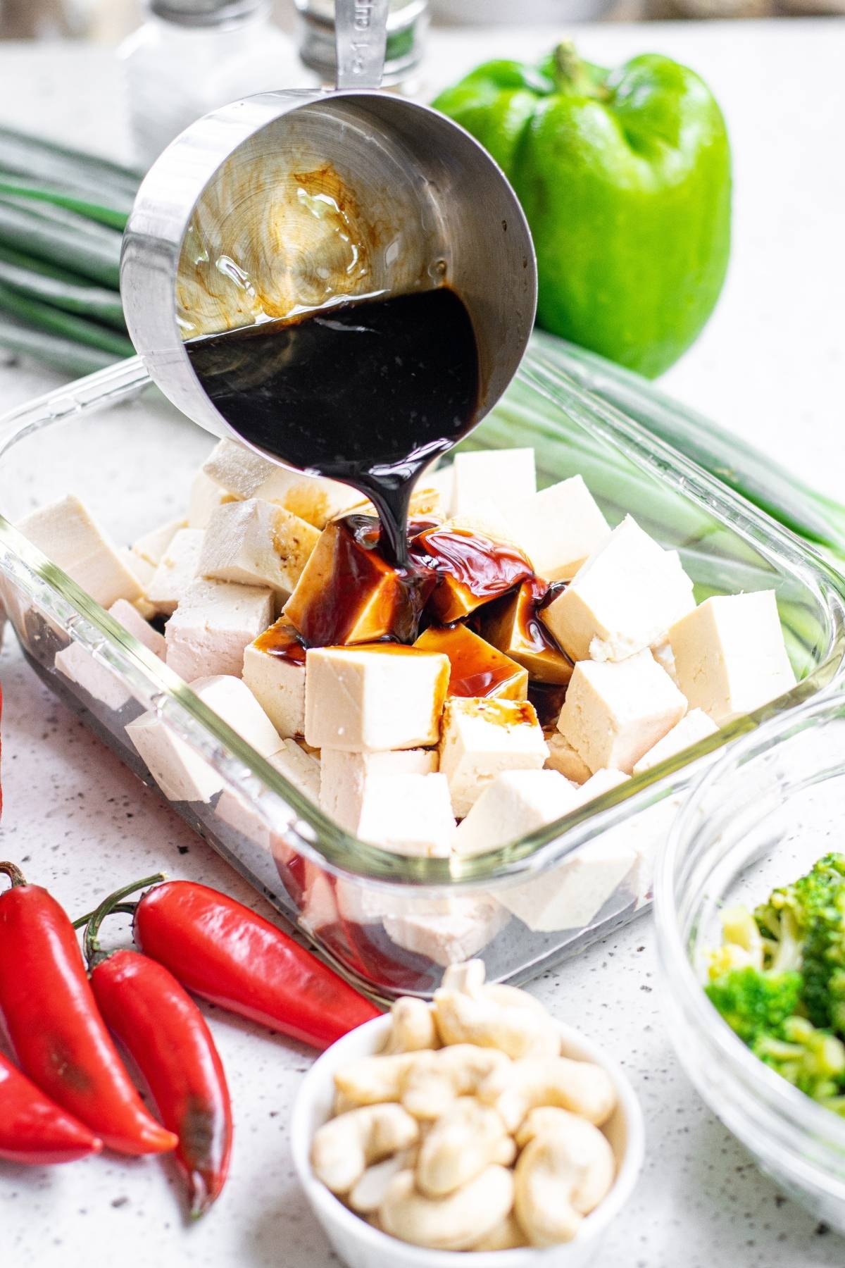 Soy sauce being poured over cubed tofu in a glass dish, surrounded by fresh vegetables and nuts—a delicious vegetarian Chinese inspired dinner perfect for any night.