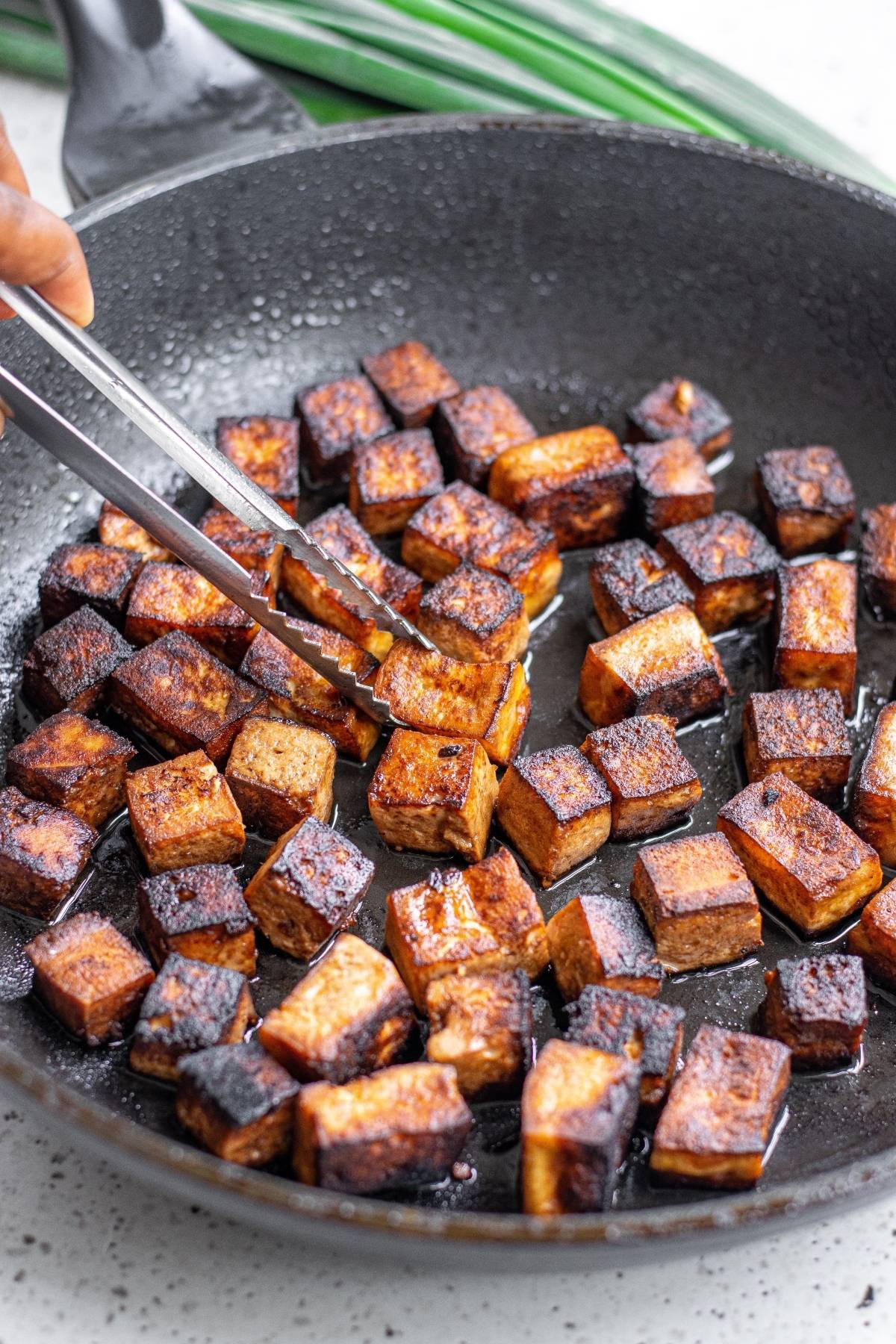 Crispy, browned tofu cubes being cooked in a black skillet, held by metal tongs—a delicious start to a Vegetarian Chinese inspired dinner or your favorite Kung Pao Tofu recipe.