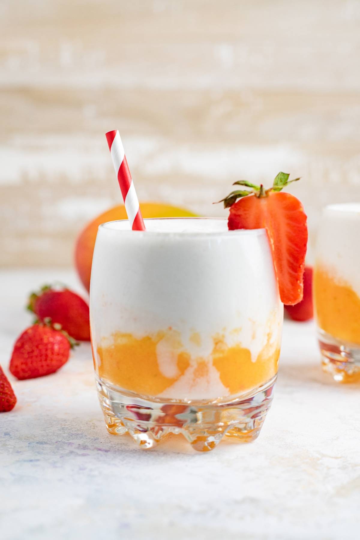 A creamy orange and white tropical rum cocktail is served in a glass with a red-striped straw and a strawberry garnish.