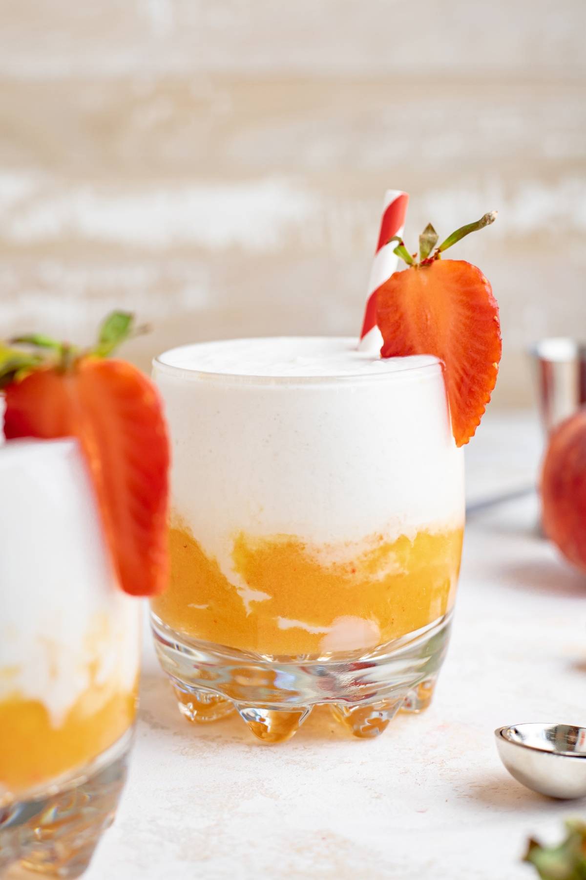 A layered mango colada slushie in a glass, blending peach and cream, garnished with a strawberry and a red striped straw.