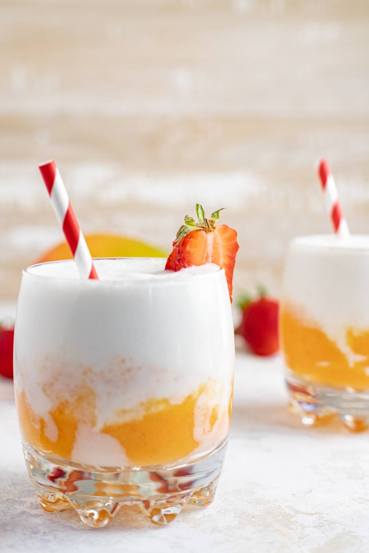 A layered slushie drink recipe featuring mango and cream in a glass, garnished with strawberries and a red-striped straw.