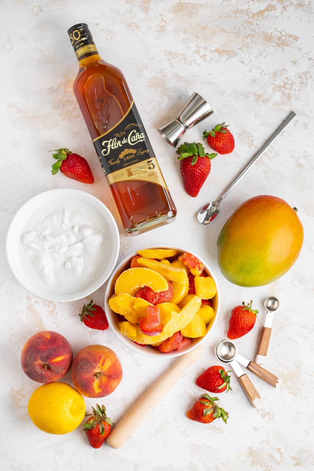 Overhead view of rum, strawberries, peach slices, mango, lemons, coconut cream, and bar tools on a white surface—perfect inspiration for a tropical rum cocktail or layered slushie drink recipe.