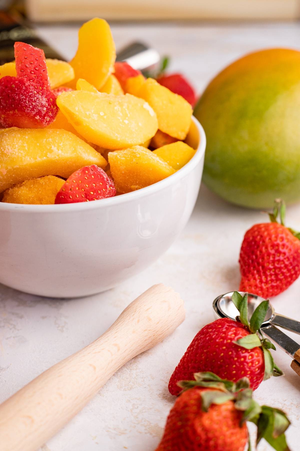 A white bowl of frozen mango slices and strawberries, with fresh strawberries and a mango nearby on the table—perfect ingredients for a vibrant layered slushie drink recipe.