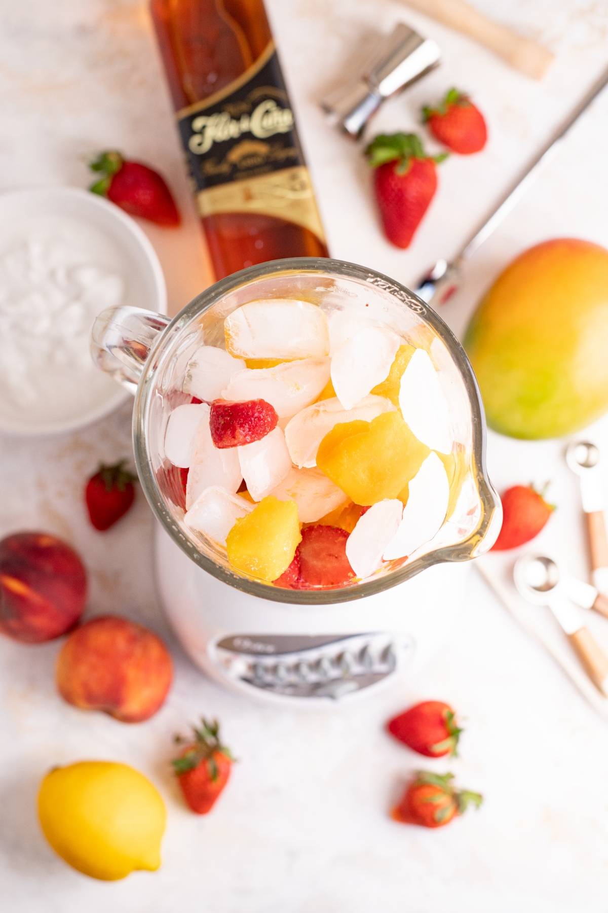 A blender filled with ice, strawberries, and mango, surrounded by fruit, rum, and cocktail tools on a white surface—perfect for crafting a Tropical rum cocktail or a vibrant layered mango colada slushie.