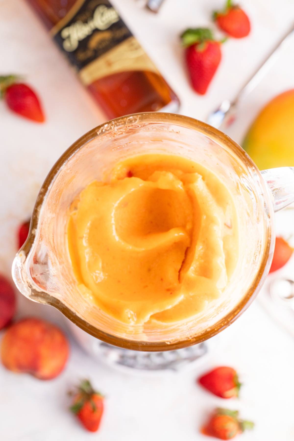 Blender filled with creamy orange smoothie, surrounded by strawberries, peach, and a bottle of liquor—perfect for whipping up a tropical rum cocktail.