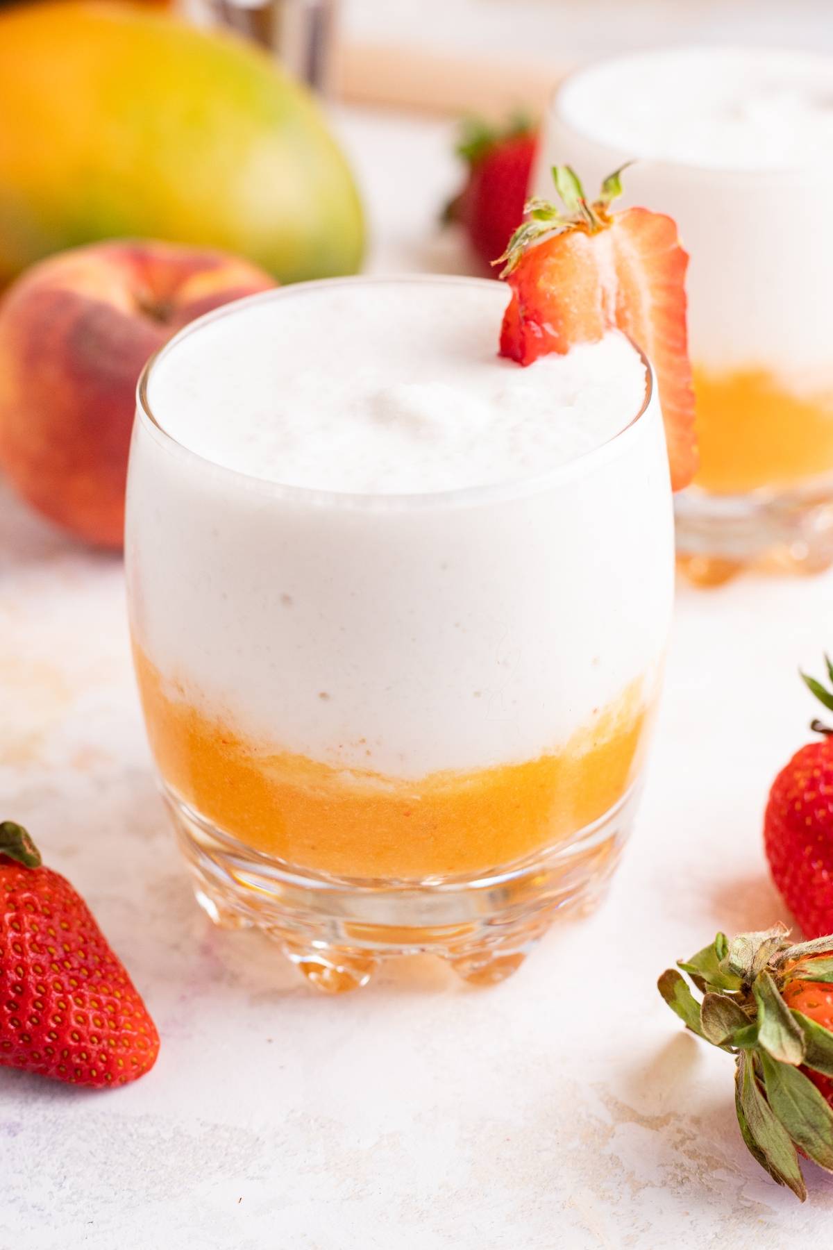 A layered mango colada slushie with white and orange sections, garnished with a strawberry slice on the rim.