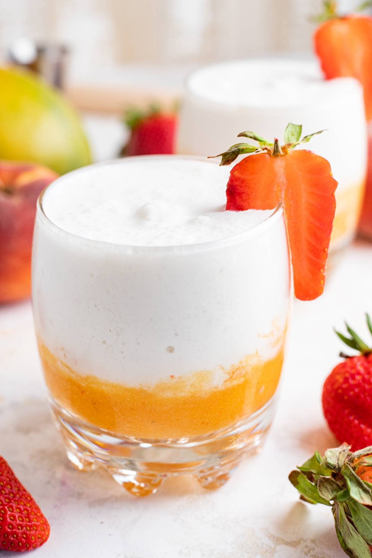 A layered mango and cream smoothie in a glass, garnished with a sliced strawberry on the rim, reminiscent of a refreshing layered mango colada slushie.