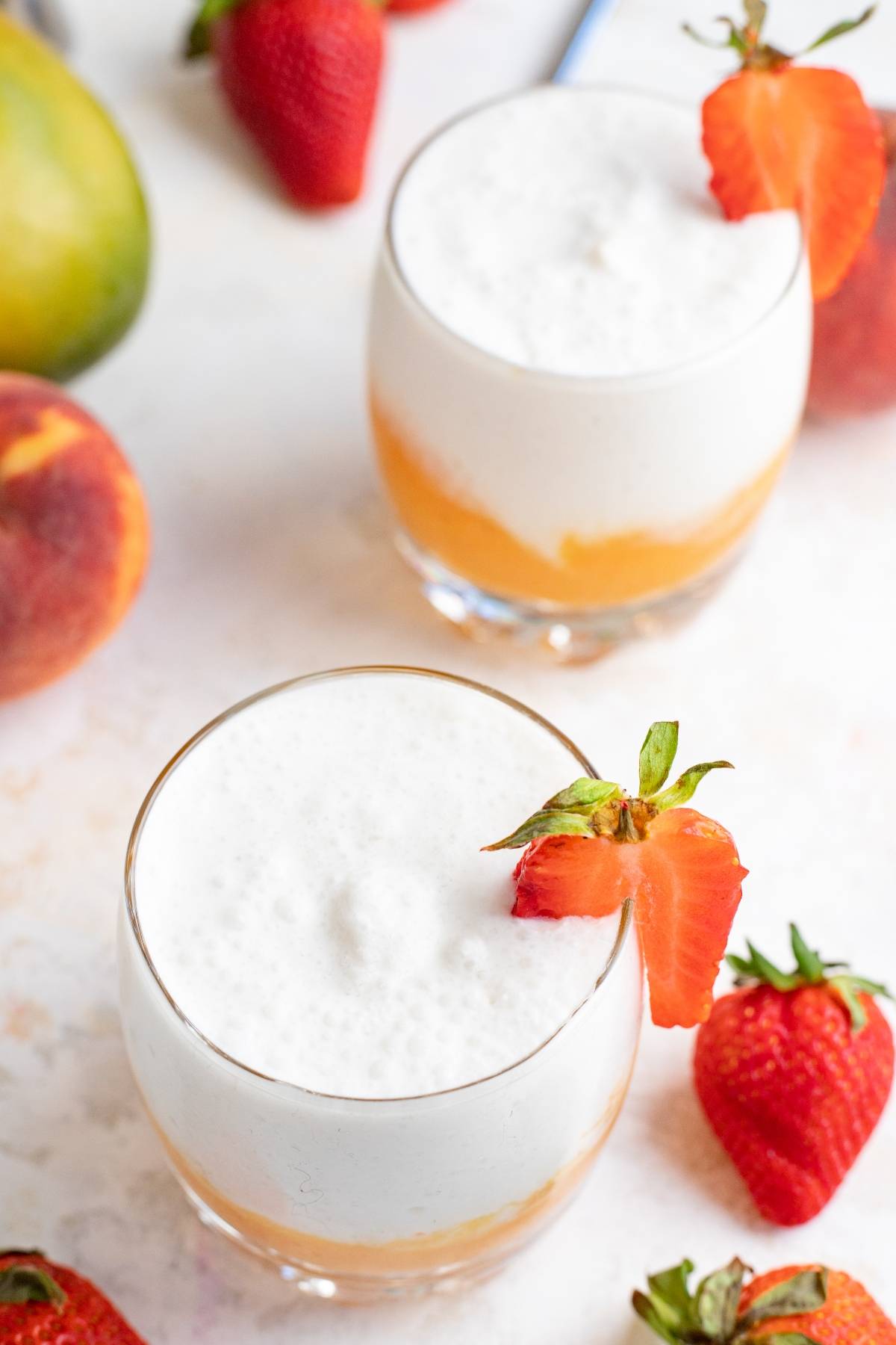 Two glasses of frothy white drinks with orange layers, garnished with fresh strawberries and surrounded by fruit—a vibrant tropical rum cocktail that's as stunning as it is delicious.