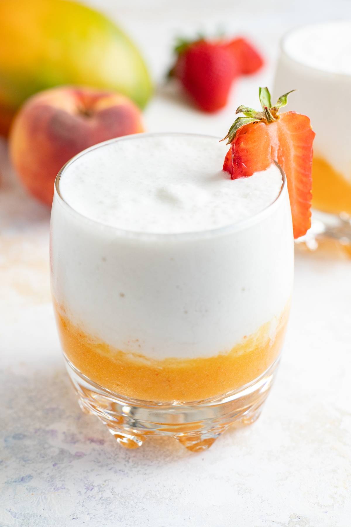A layered peach and cream smoothie in a glass, garnished with a strawberry slice, reminiscent of a layered mango colada slushie.